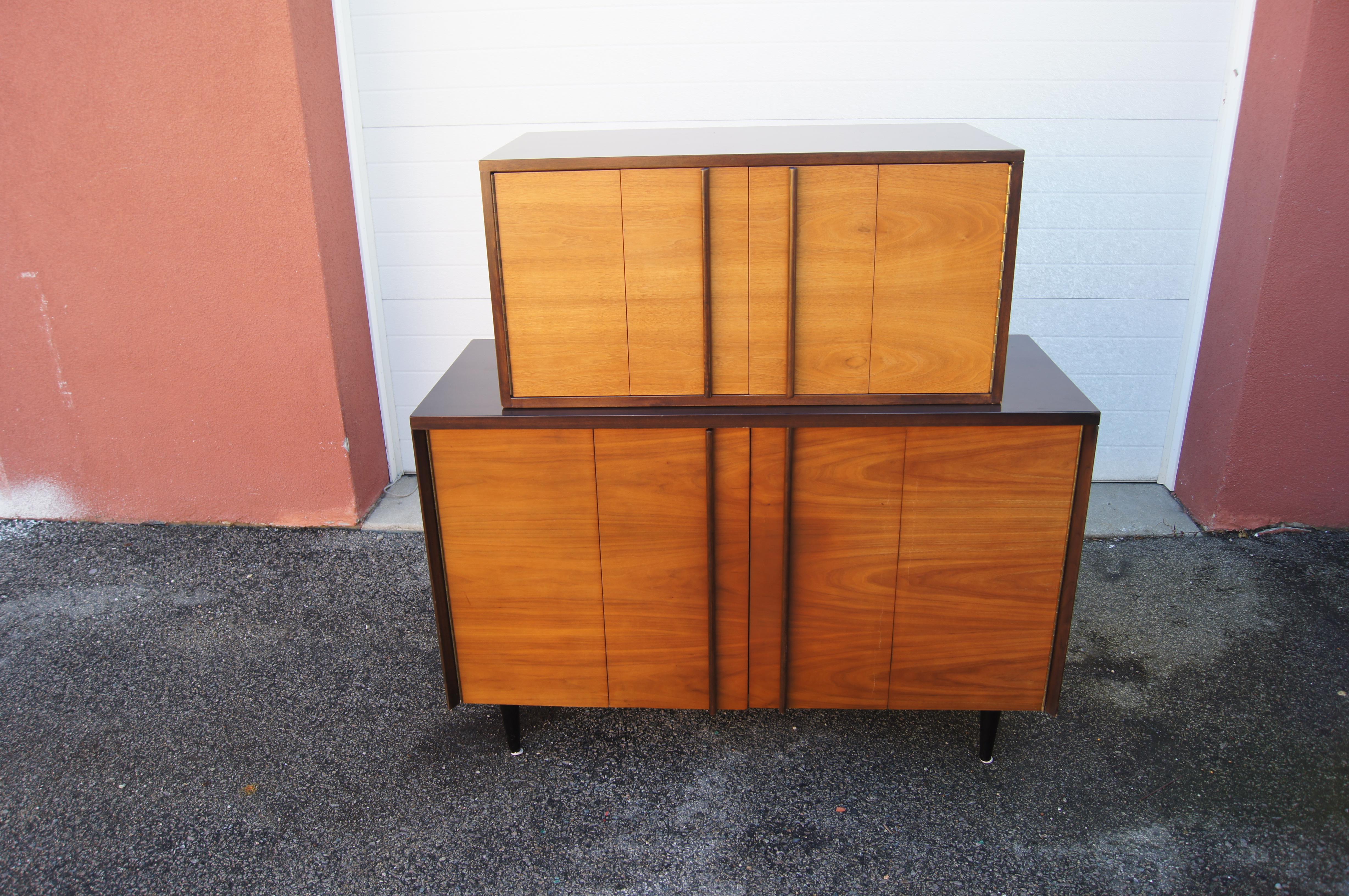 Designed by John Stuart, this mid-century highboy dresser comprises two walnut cases whose dark frames and vertical door pulls contrast with the warm-toned fronts. The top case contains three wide drawers, one of which is divided in half and one of