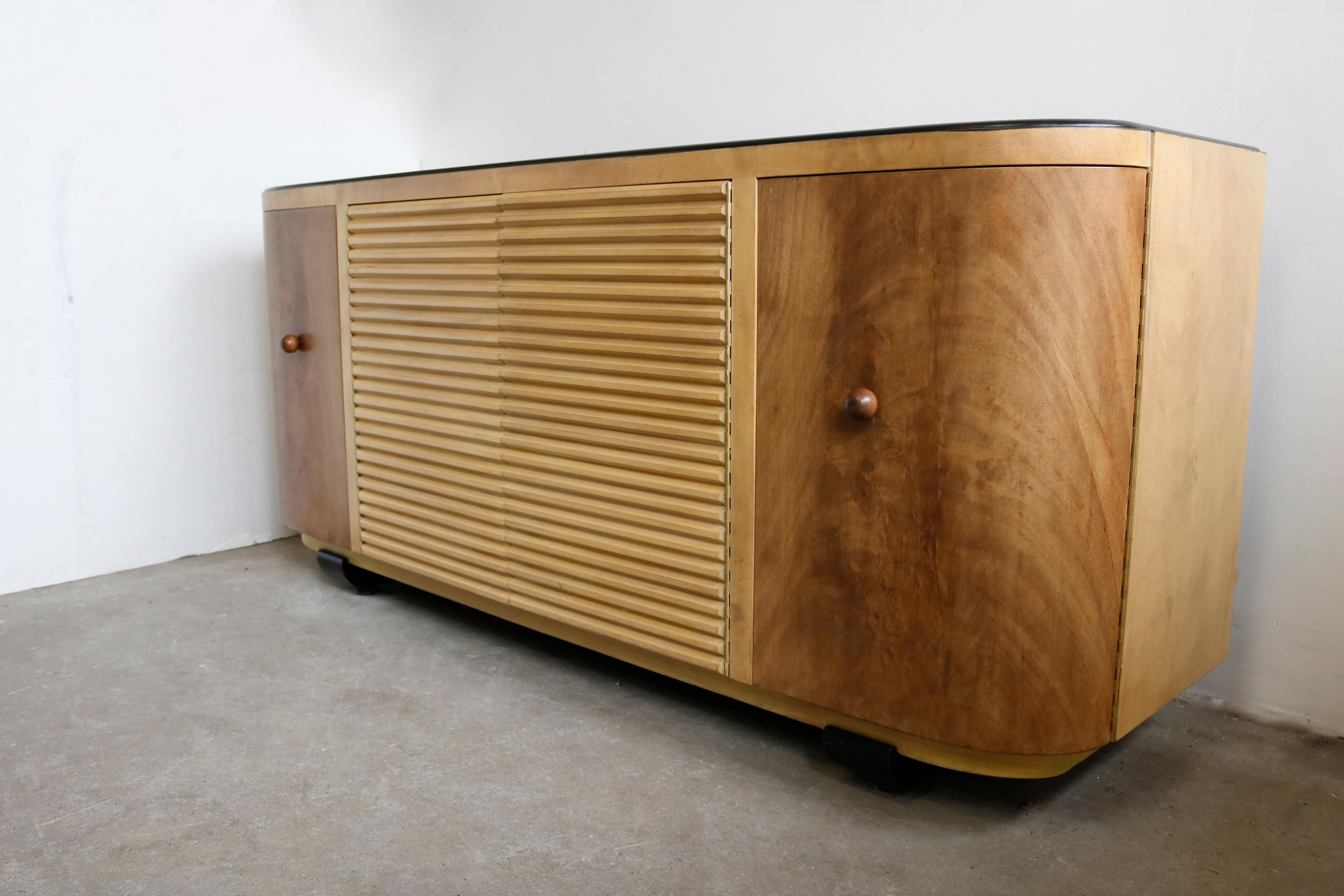 A modern, two-toned credenza in flame mahogany and sycamore with a smooth ebony finish top and legs by Paul Frankl.
The right side doors open on 1 drawer and a shelf and the left one on 4 with a rounded front.
The 2 middle push-to-open slatted