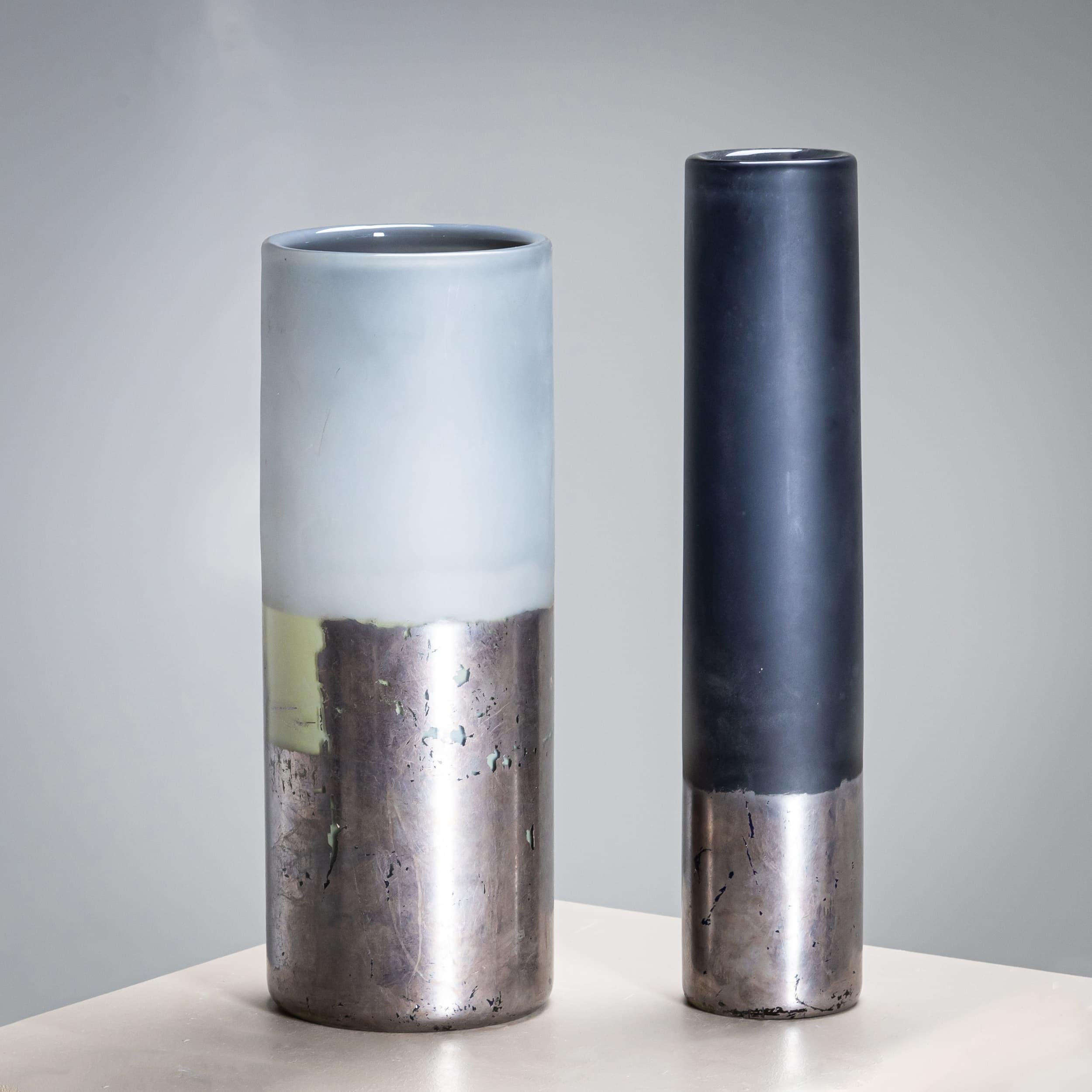 Coppia di vasi in vetro dalle forme cilindriche leggermente diverse (H: 29 x Ø6 cm e H: 27 x Ø10 cm), nei colori antracite e grigio chiaro. Il terzo inferiore è patinato in argento. La base reca una firma illeggibile e la dicitura 