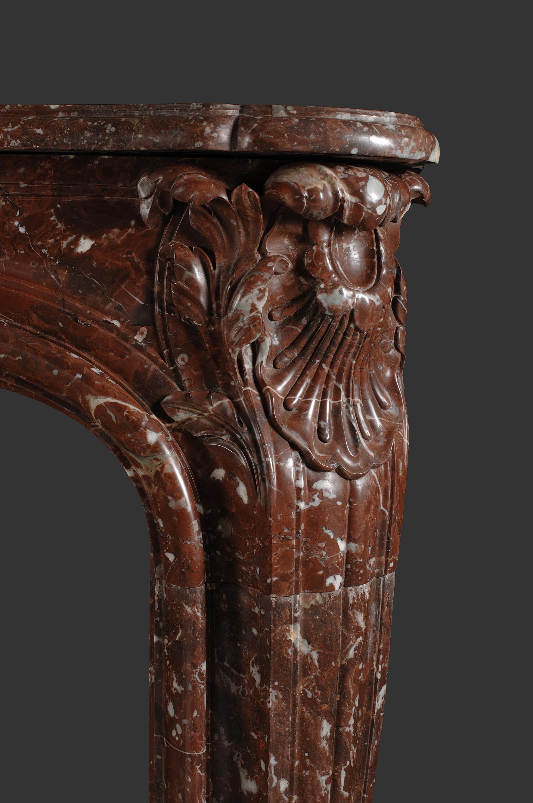 Ein Louis XV Kamin mit Serpentinenregal aus altem belgischem Griotte-Marmor. 

Der Fries ist mit der für Ludwig XV. typischen symmetrischen Kartusche verziert, die von schön geschnitzten Blumen flankiert wird,  Die Endblöcke des Frieses sind mit