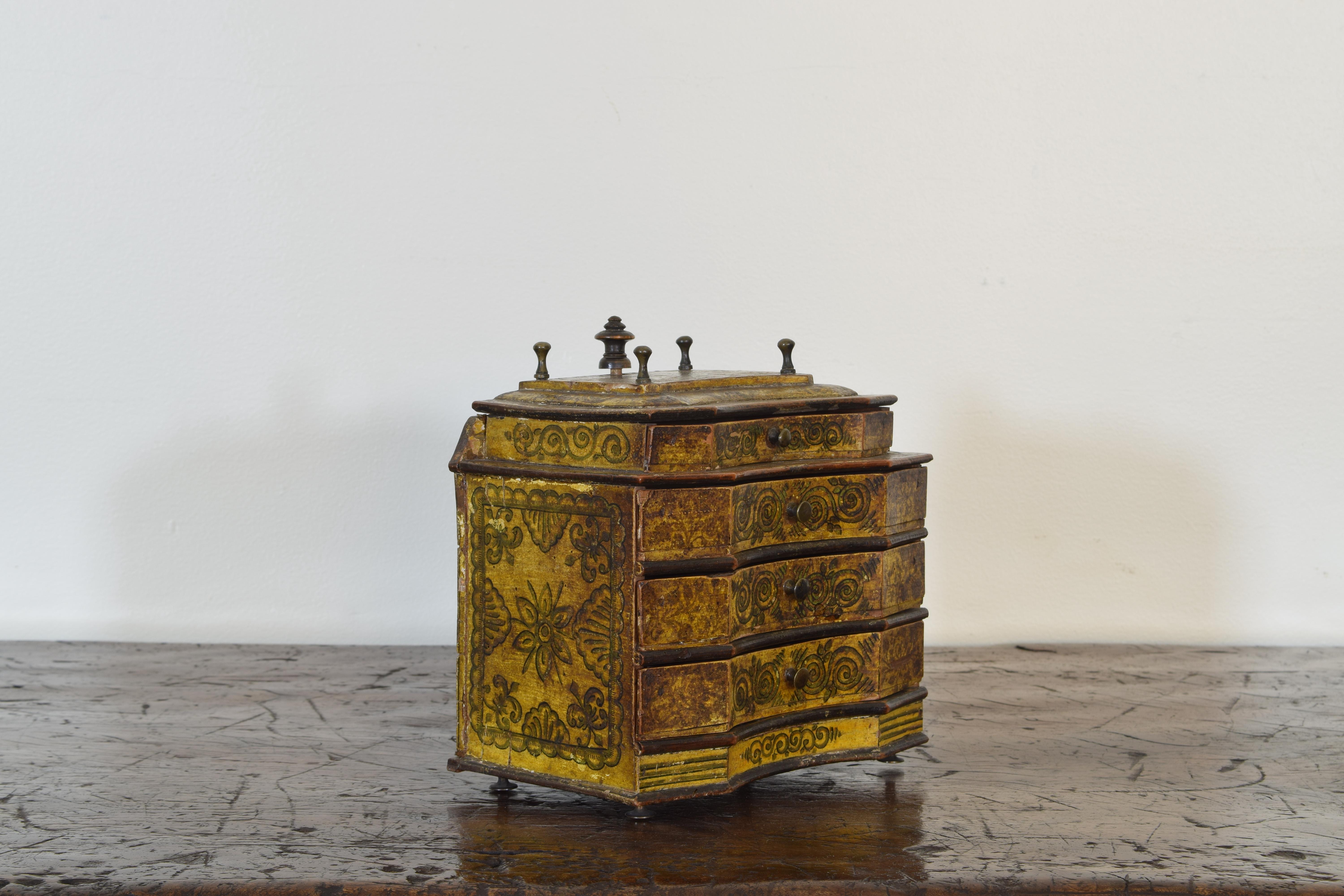 The box with original painted and decoration having brass finials and a larger wooden finial that is the locking mechanism for all of the drawers, the ochre colored paint further decorated with scrolls, foliage, and shells, raised on small brass