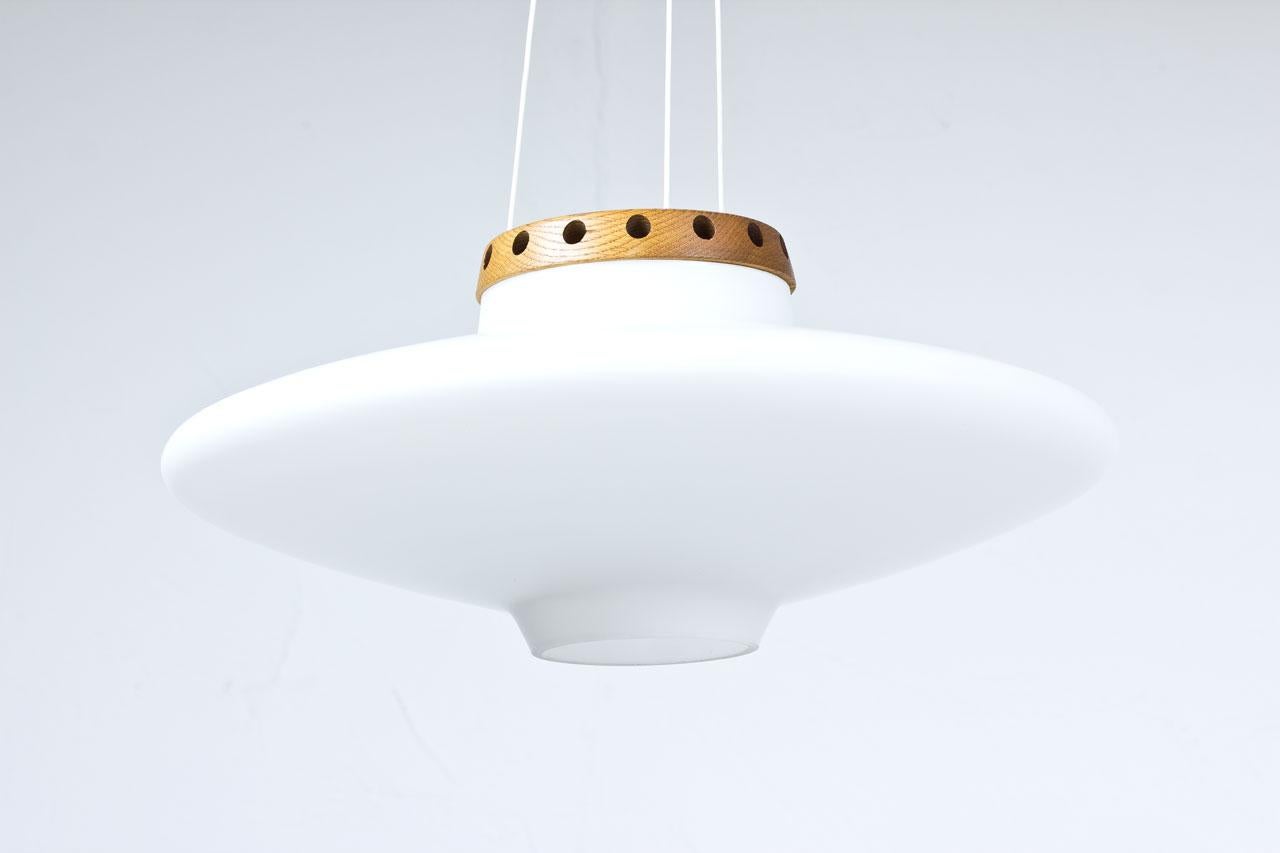 UFO pendant lamp model 565 by Uno & Östen Kristiansson. Manufactured by their own company Luxus, at Vittsjö in Sweden during the 1950s. Made from opaline glass diffuser with
solid oak fittings.
