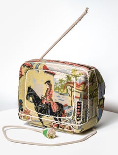 "TV", donna a cavallo a punto croce avvolta da un televisore d'epoca con presa di corrente