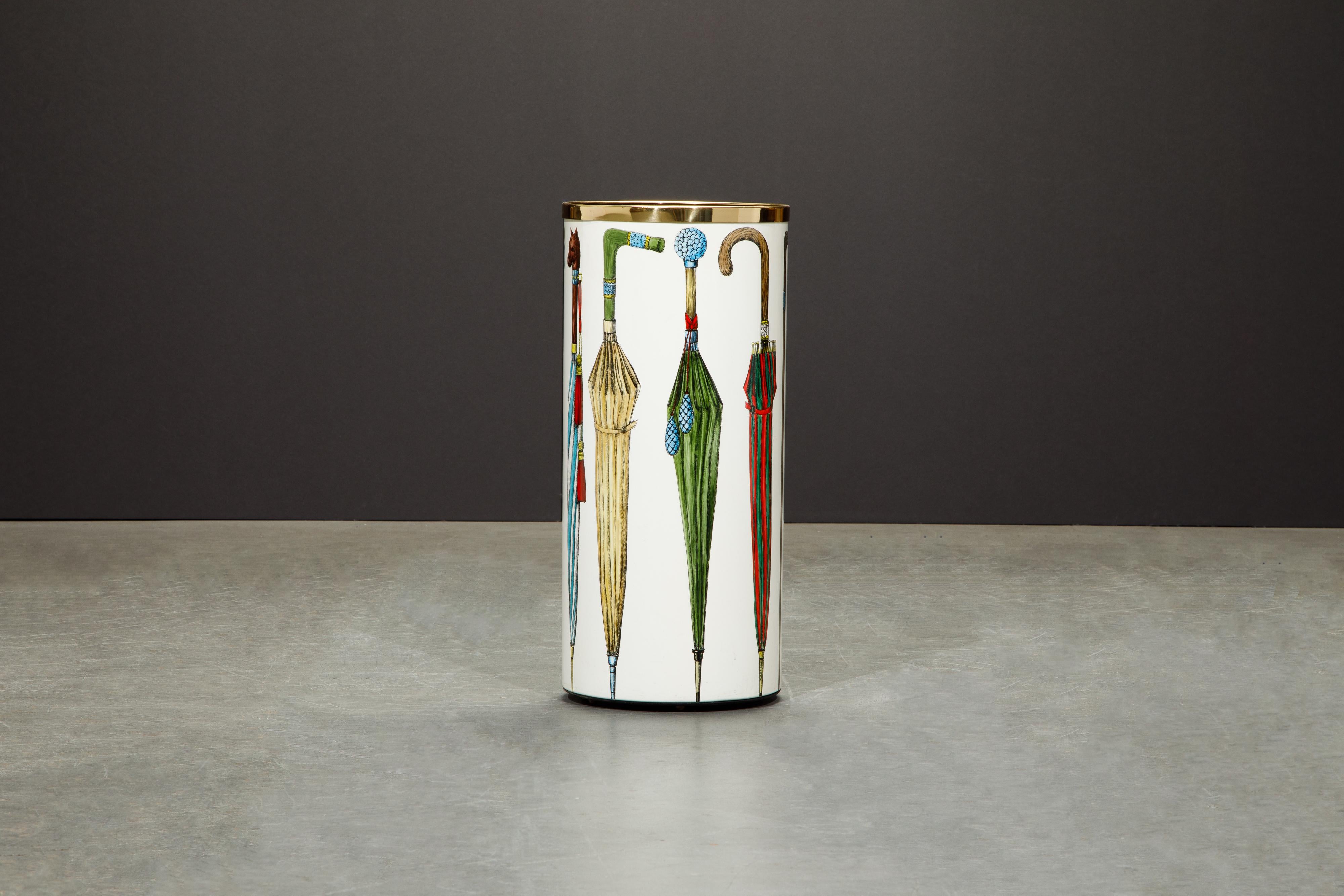 This umbrella stand is by Piero Fornasetti, signed underneath with label. This round umbrella bin is made with lithographic transfer and lacquered metal that has a white background with colorful umbrella motif with brass trim and brass feet.