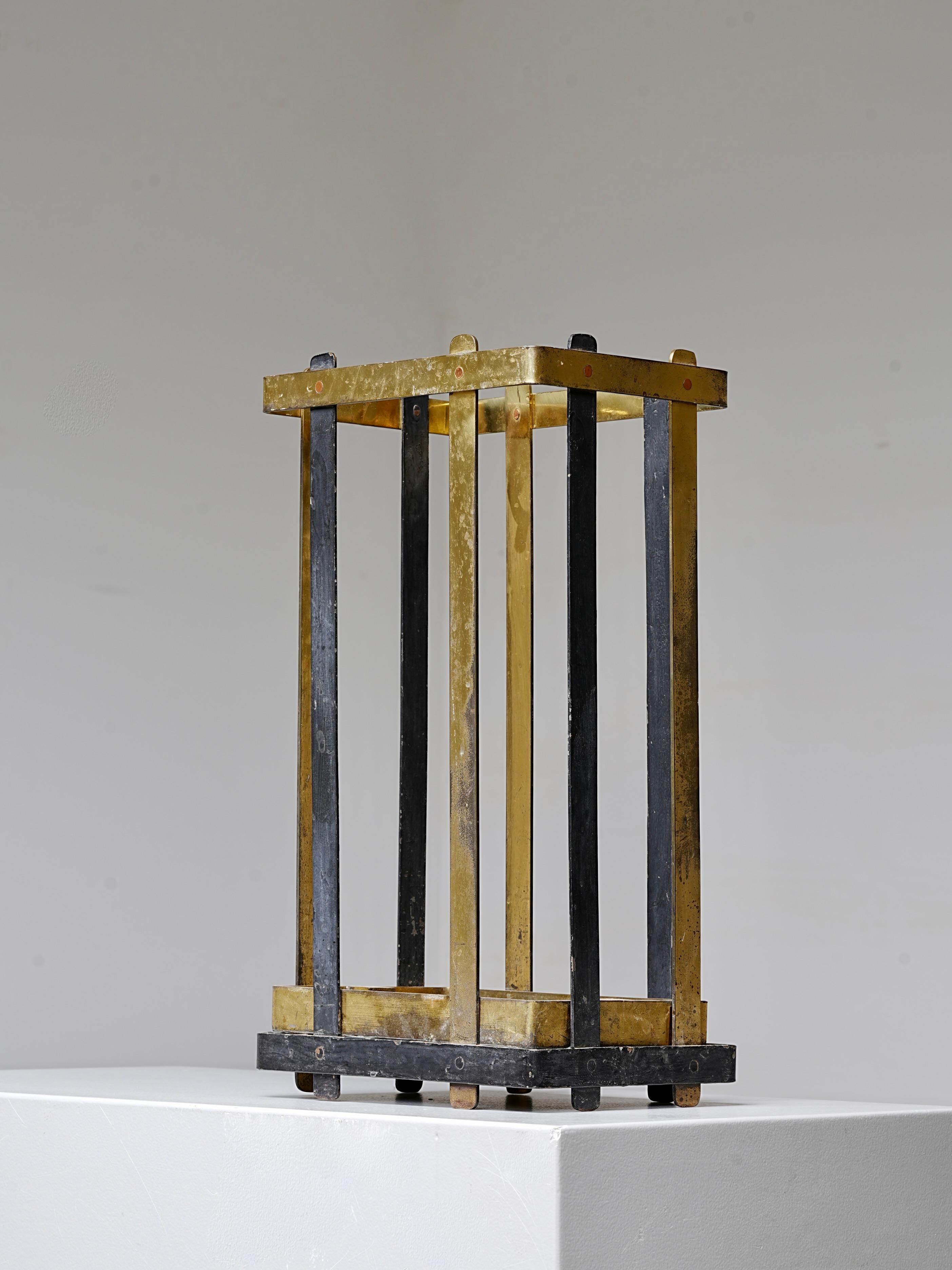 Dieser Schirmständer kombiniert Metall und Messing auf eine praktische und durchdachte Weise. Der schwarze Stahlrahmen verleiht dem Möbelstück eine klare, minimale Struktur, während der geflochtene Rattaneinsatz Wärme und Textur verleiht. Eine