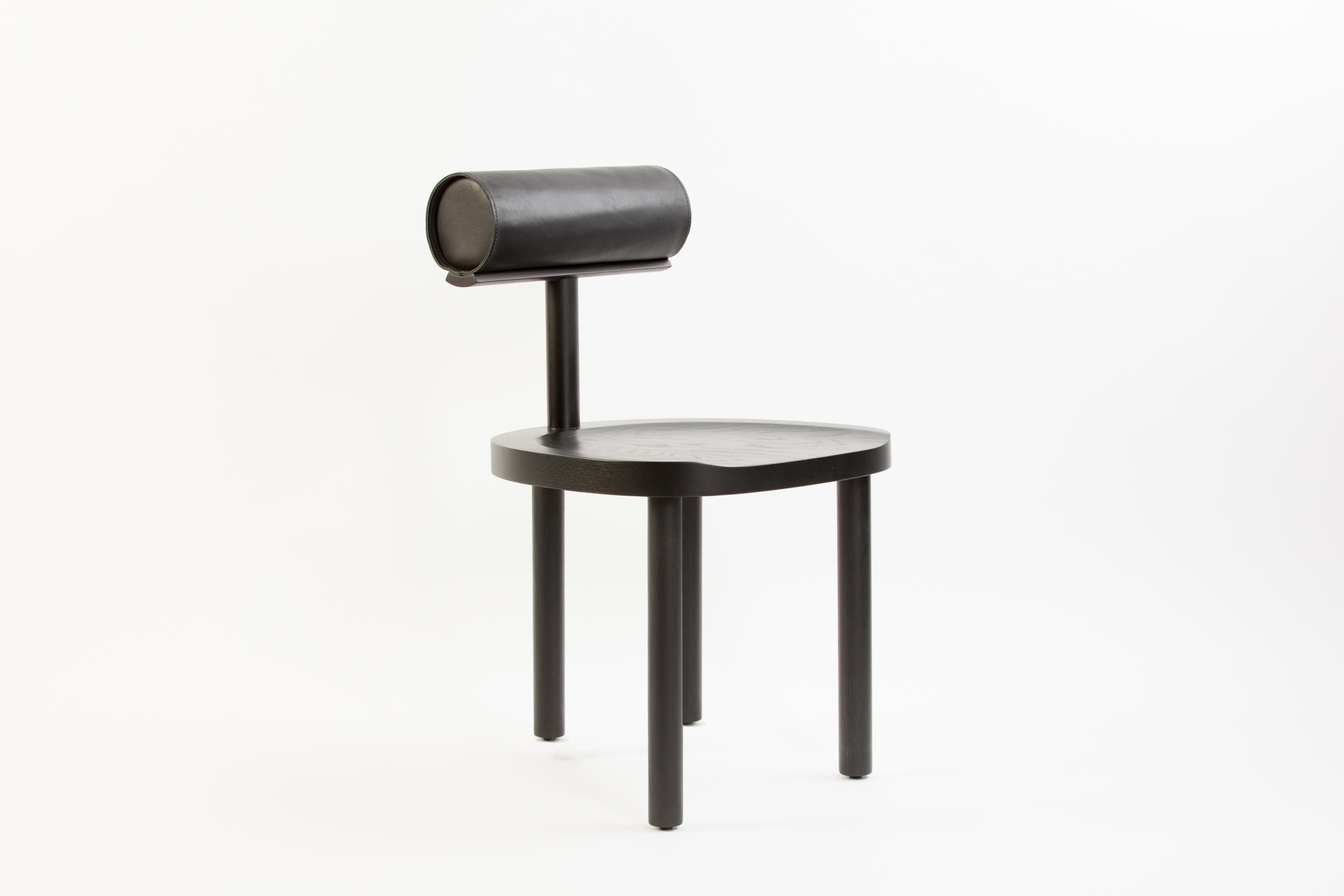 La chaise UNA croise un siège et des pieds ronds en bois avec un dossier cylindrique recouvert de cuir. L'utilisation de ces formes fluides permet de mettre davantage l'accent sur les détails du grain du bois et du dessus en cuir cousu à la