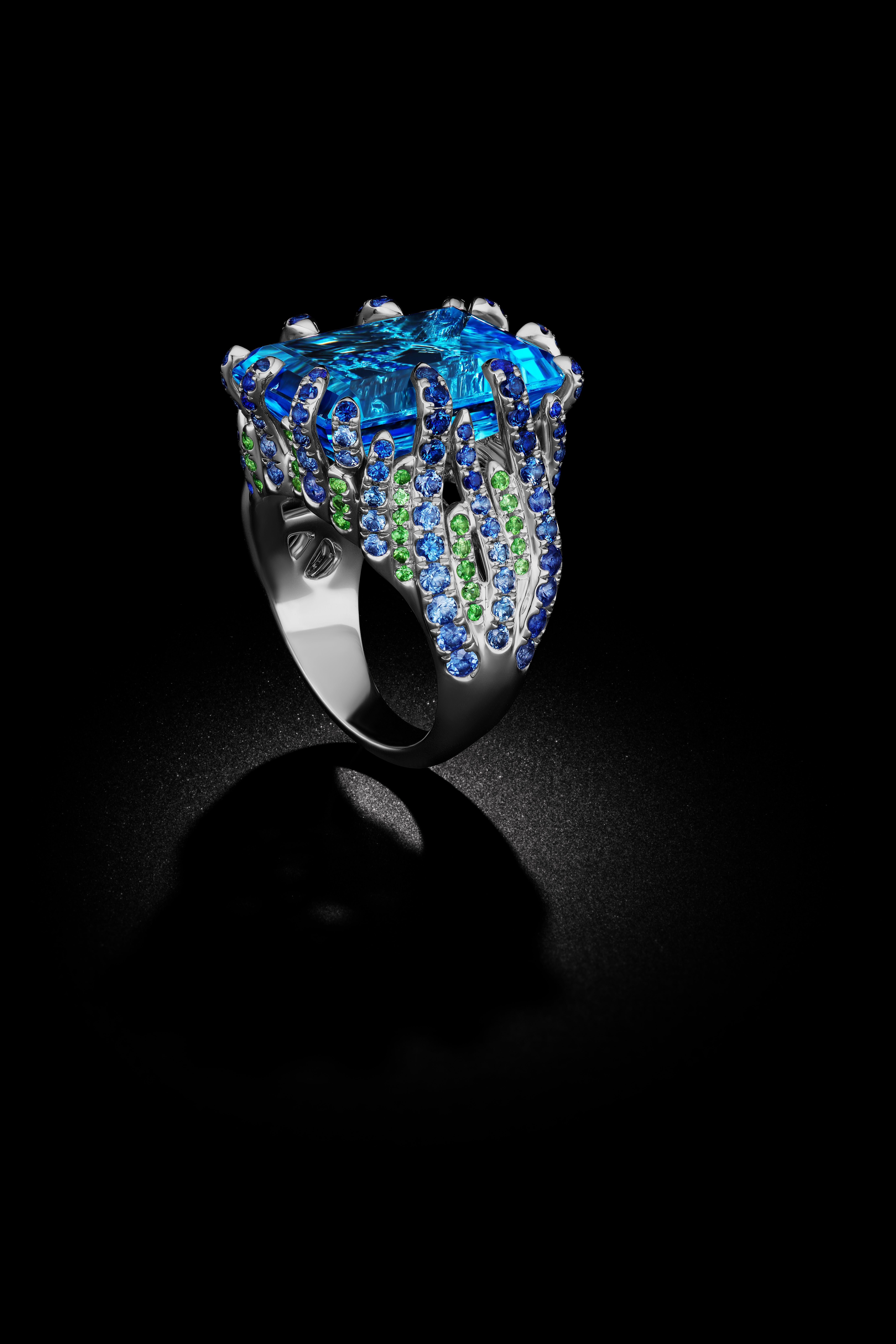 The center of the Blue Topaz  gemstone bears a resemblance to the deep blue water of the sea. A look at the ring's side view are pave of Tsavorites (Green Garnets) and dark Sapphires creating the the illusion of waving fronds in the sea. This