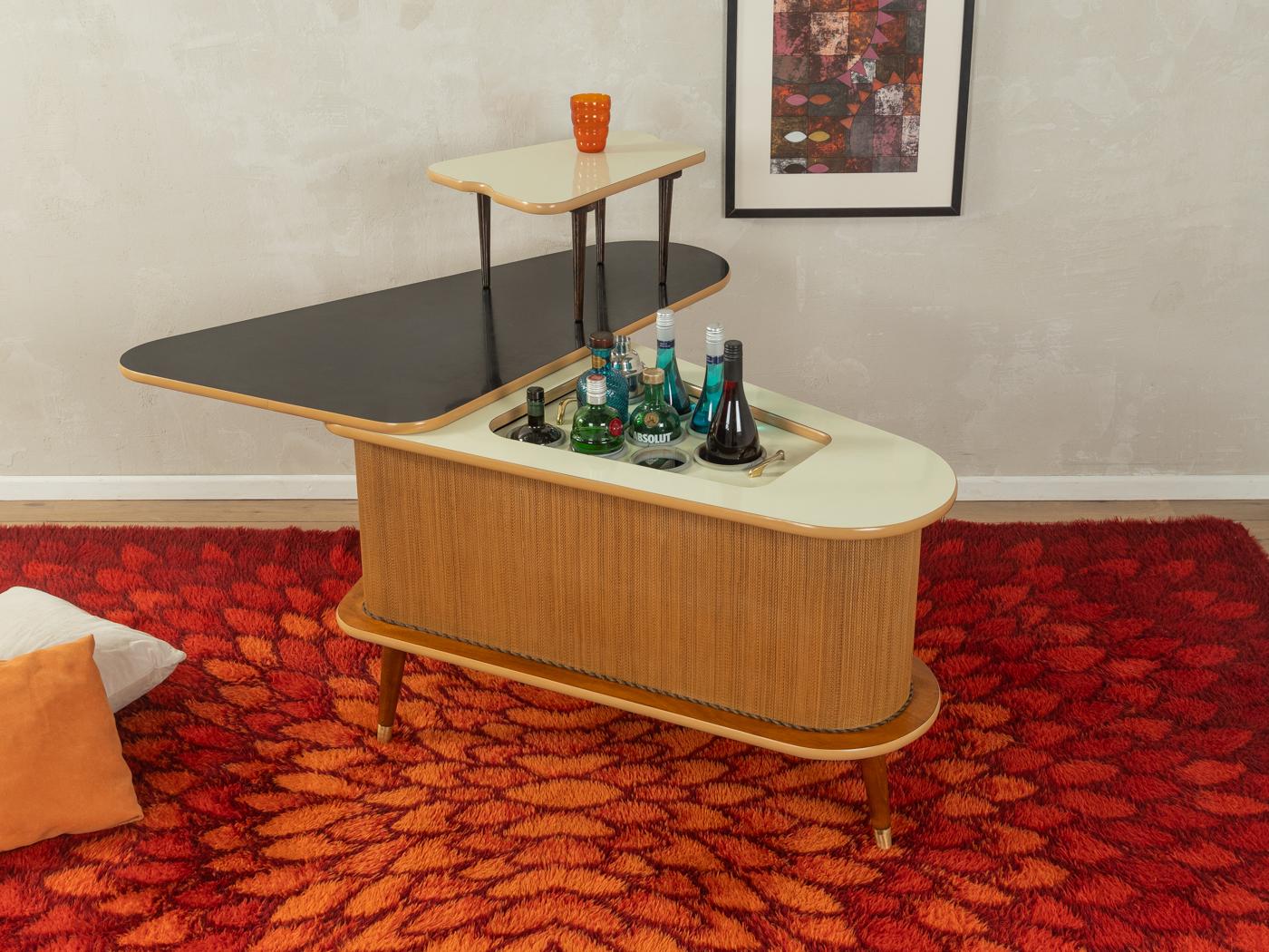 Bar unique datant des années 1950. Corpus avec galon en rotin d'origine, garnitures décoratives, un compartiment bar avec porte-verres et porte-bouteilles, une table d'appoint amovible et des pieds inclinés avec capuchons en laiton. Surfaces avec