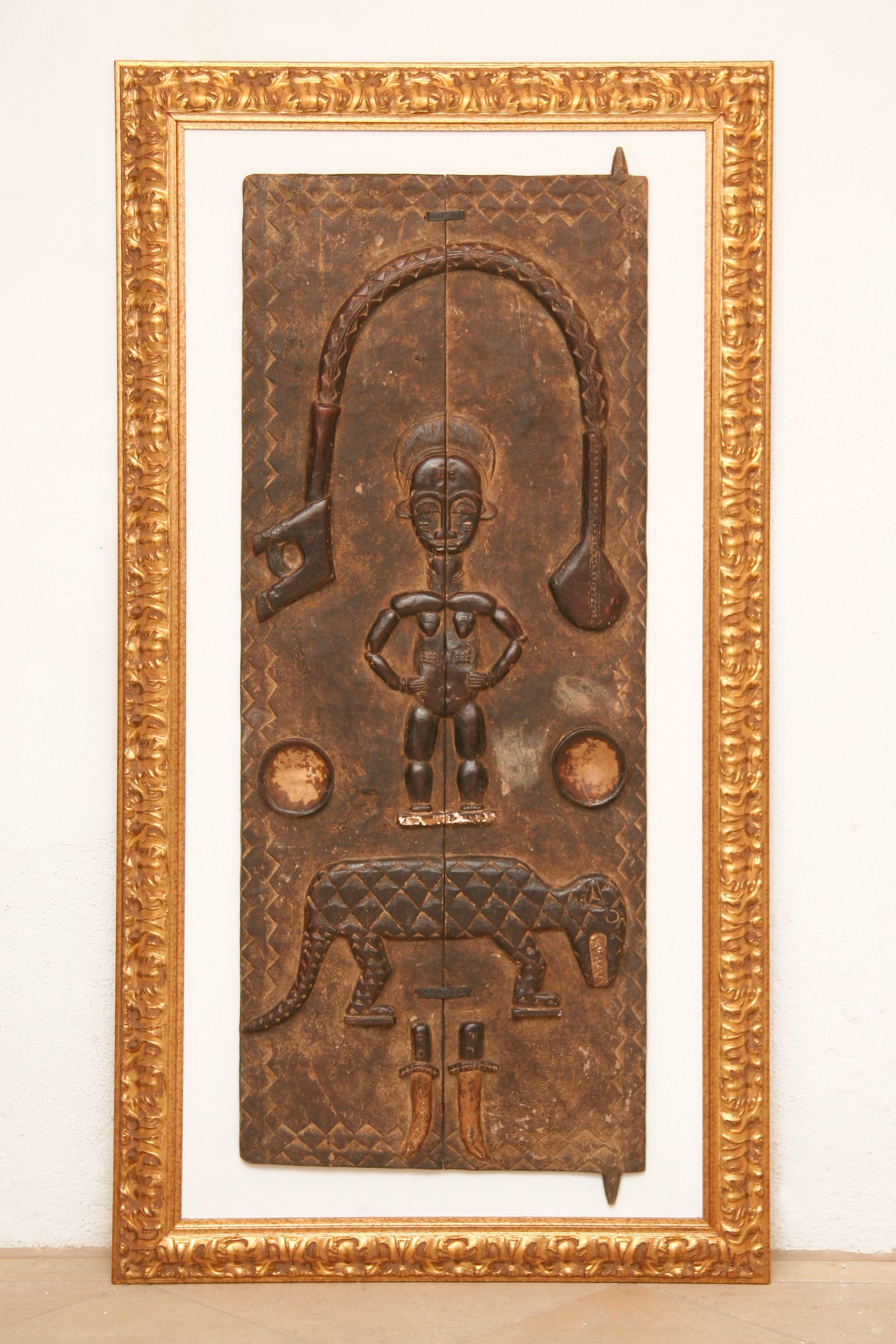 Portes en bois massif sculptées à la main d'une armoire africaine (Mali/Dogon). 
fixée sur une plaque de bois de couleur blanche, encadrée d'or.
L'objet présente des symboles africains typiques : 1 figurine, 1 arme, 1 léopard, 1 couteau. 
qui, en