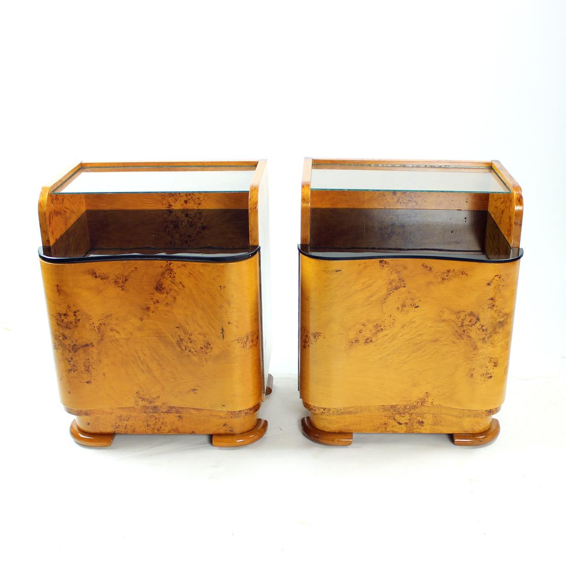 Unique set of two bedside tables from art deco era. Produced in Czechoslovakia in 1940s. The tables are made of oak wood with walnut veneer finish. Each table has a compartment with doors and one glass shelf in clear glass. There is also a black