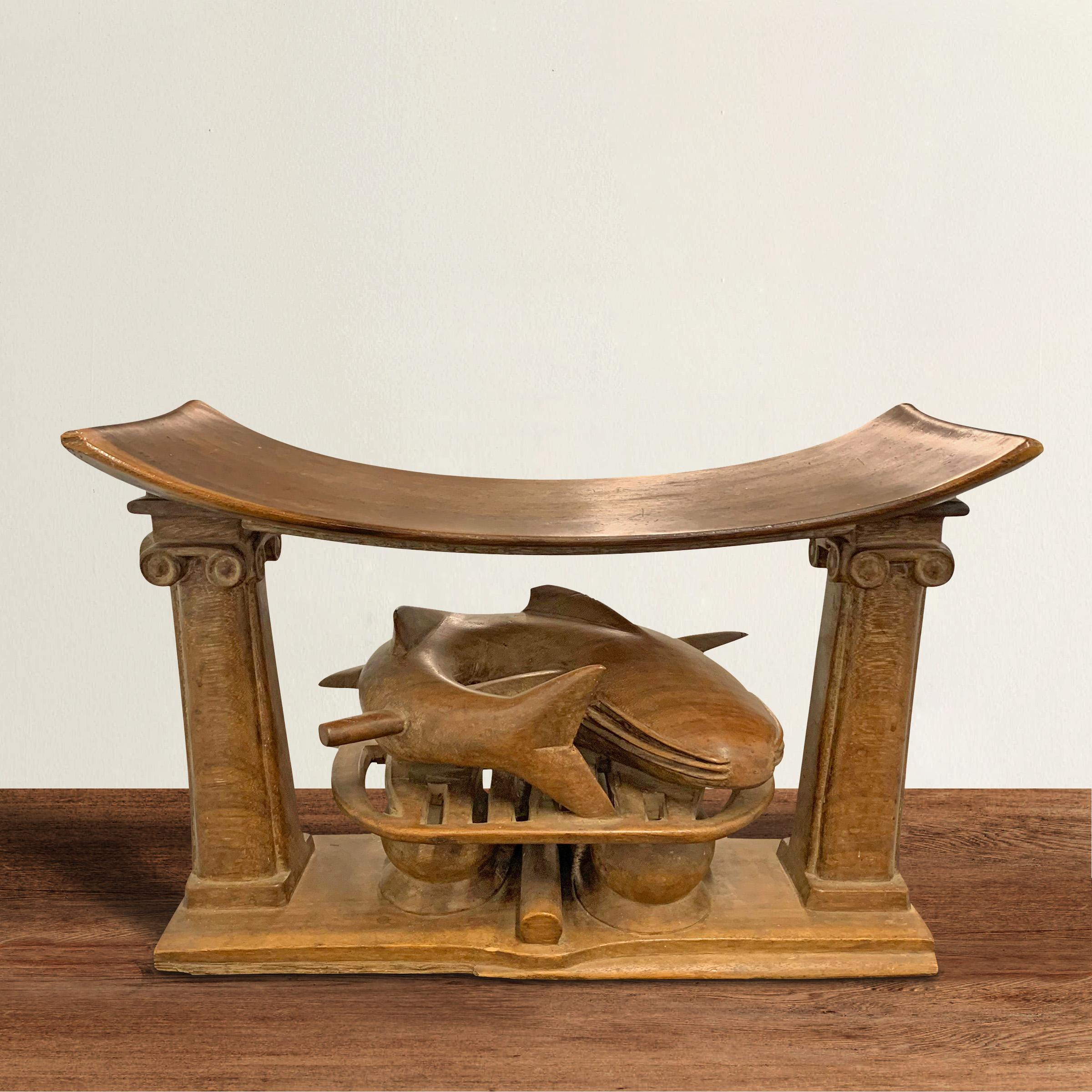 A unique Asante stool depicting a speared catfish on a grill resting under a a curved seat supported by square ionic columns, and all carved of one piece of wood.