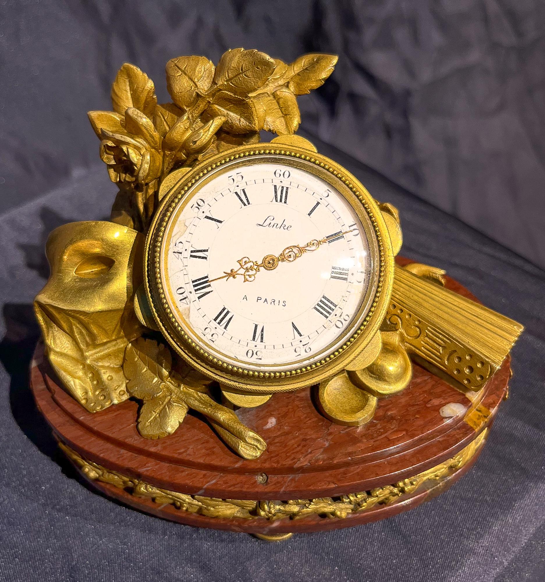 This exquisite Linke desk clock's face is nestled within a tamborine, that rests on a bouquet of roses, a masquerade mask, and a fan. The collection of items undoubtedly references a musical or the aftermath of a grand 18th century ball. Clocks from
