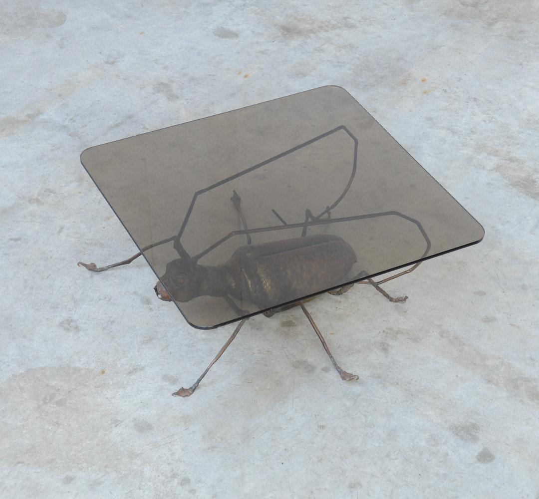 Cette table basse décorative peut être datée des années 1970 et est probablement fabriquée en Belgique.
La grande sculpture de grillon en métal fabriquée à la main est la base de cette table basse unique avec un plateau carré en verre