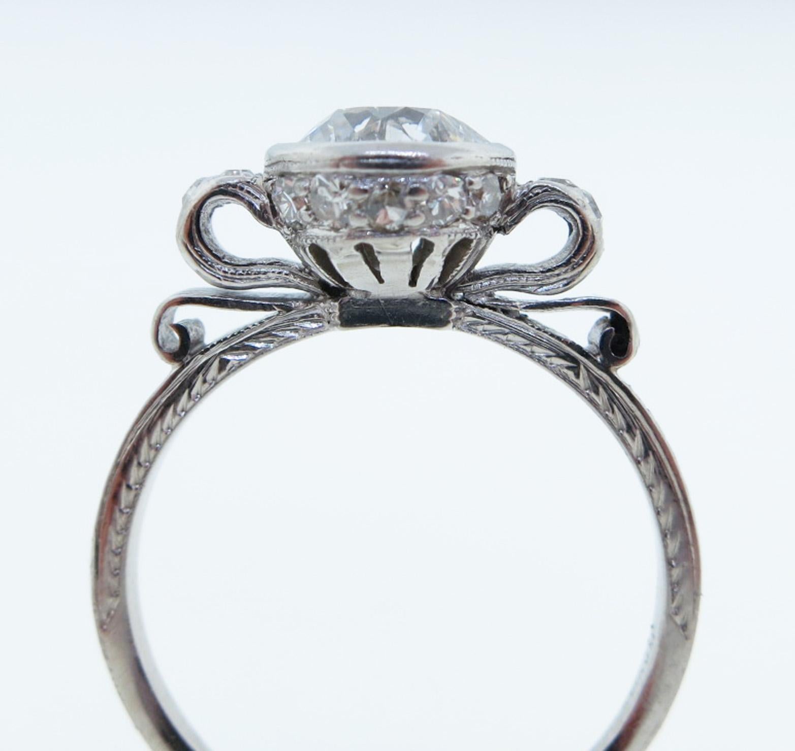 Handmade platinum mount diamond ring in a bow design mount. The center is bezel set with a round brilliant cut diamond weighing approx. 1.0 cts. grading SI1 clarity I-J color, the engraved mount is bead set 14 round diamonds totaling approx. .20cts.