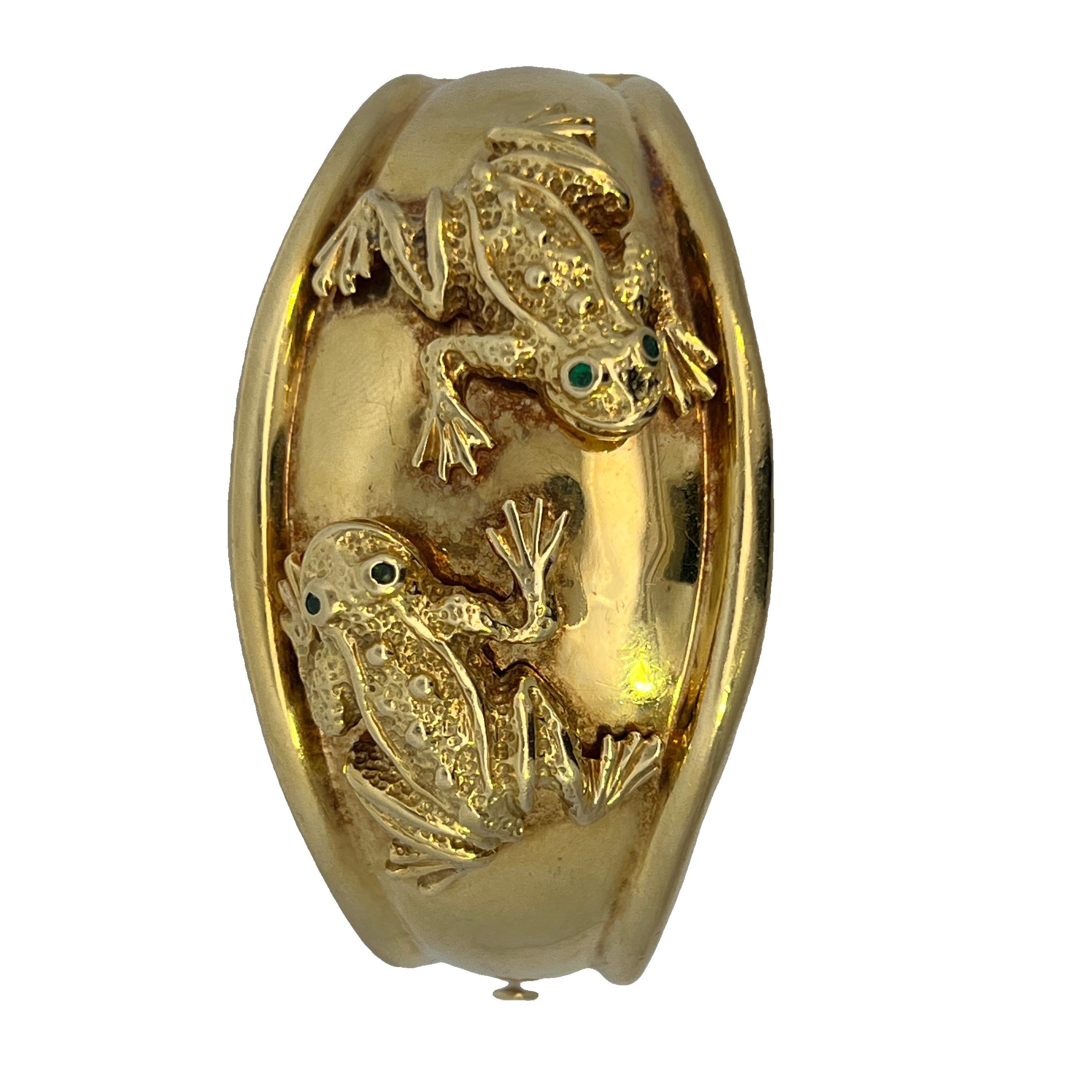 Un bracelet manchette en or magnifiquement taillé, mettant en scène deux grenouilles espiègles aux yeux d'émeraude.  Le motif présente un bord roulé de part et d'autre de la manchette, qui sert à accentuer le motif central. Le fermoir s'enclenche en