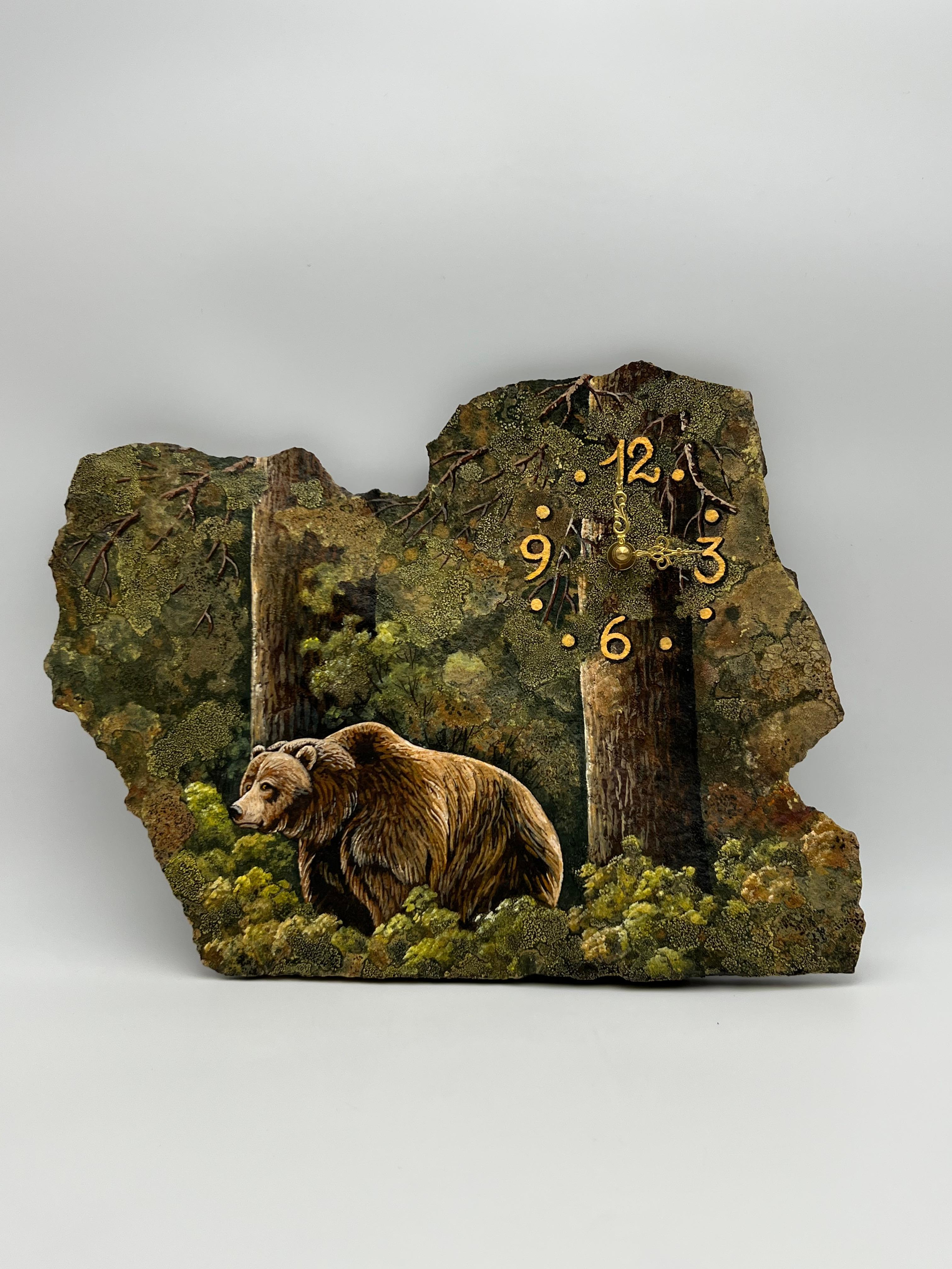 A unique sculptural wall clock crafted from a thin slice of natural stone, hand-painted with a forest scene featuring a gray bear among pine trees and grasses. The artist ingeniously incorporated the stone’s natural moss into the design, using it to