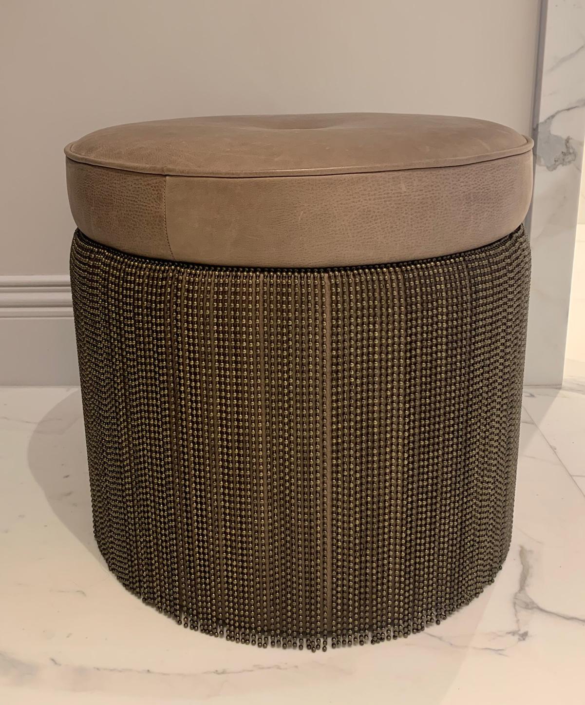 Upholstered leather ottoman. 

This contemporary ottoman features a round, soft brown leather cushion top with a brushed finish, supported by a cylindrical base adorned with antiqued bronze ball chains. The artisanal touch is evident in the unique