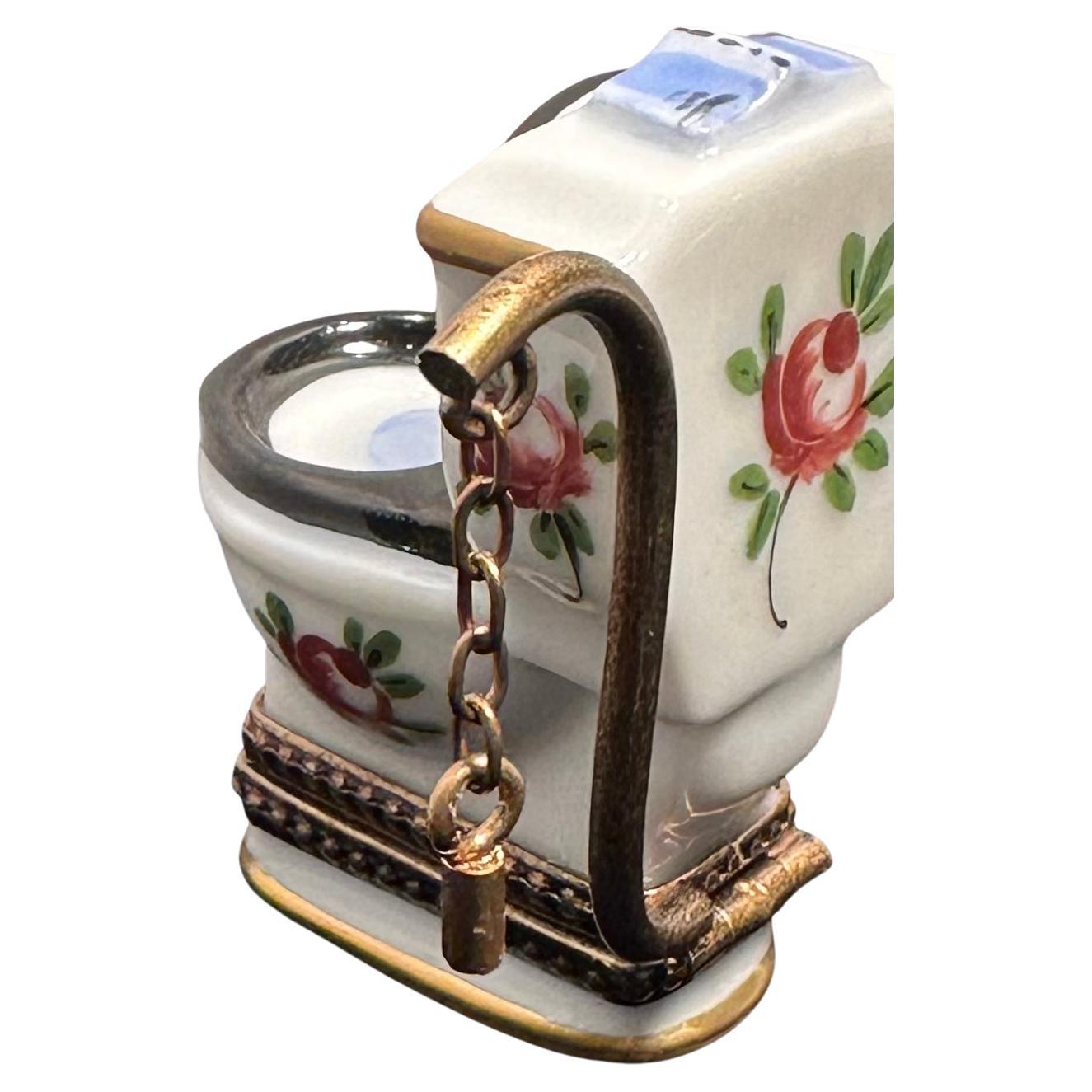 Collectible handmade and hand painted Limoges France porcelain miniature toilet shaped trinket box features a white background with pretty hand painted floral motifs accenting different areas of the toilet and a contrasting black colored seat. The