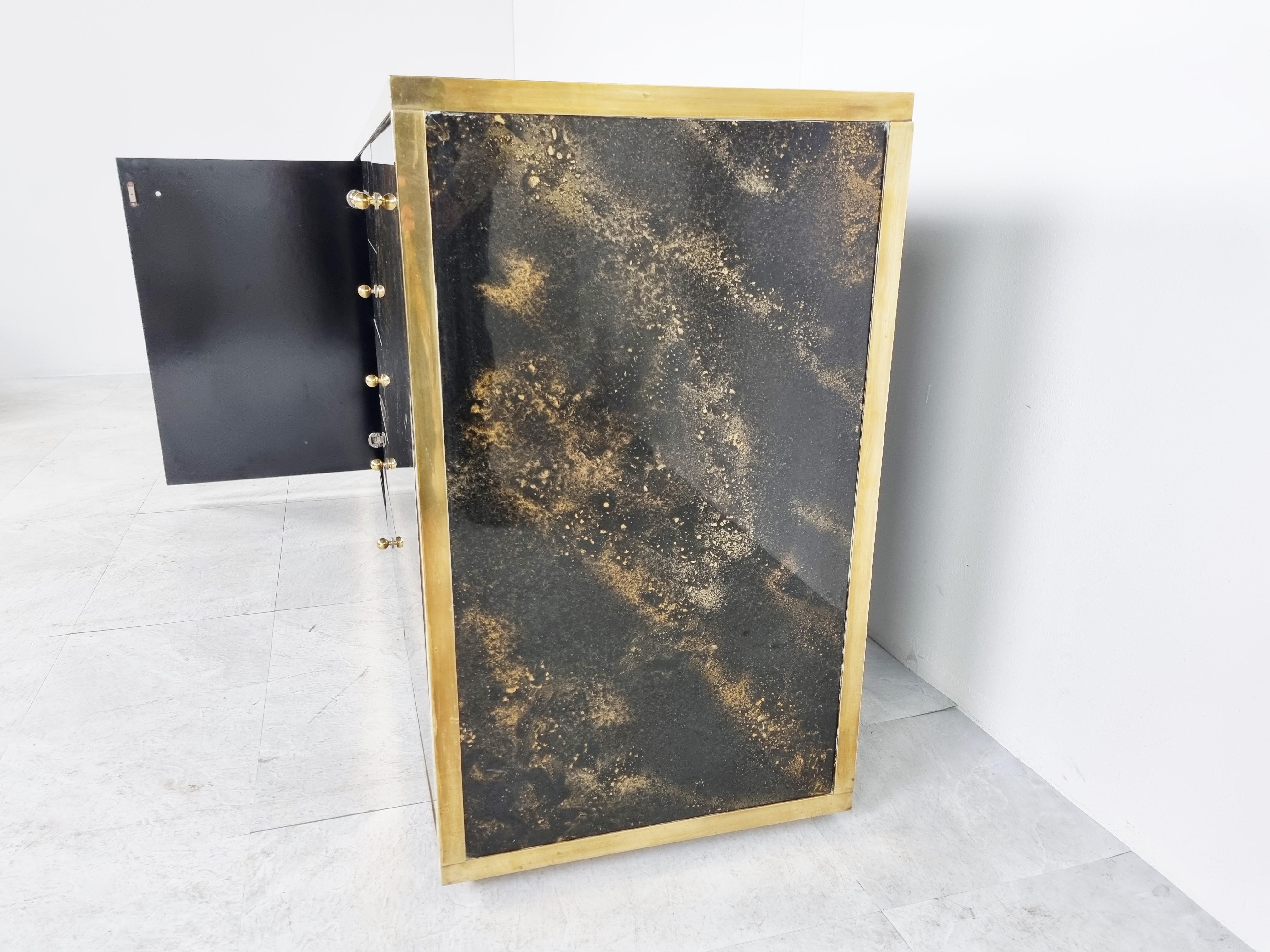 Luxurious seventies glamour sideboard by Maison Jansen consitsing of black lacquer panels with a beautiful gold flaked pattern and a patinated brass frame.

it has two doors and 5 central drawers.

Very eye ctaching piece

Good condition,