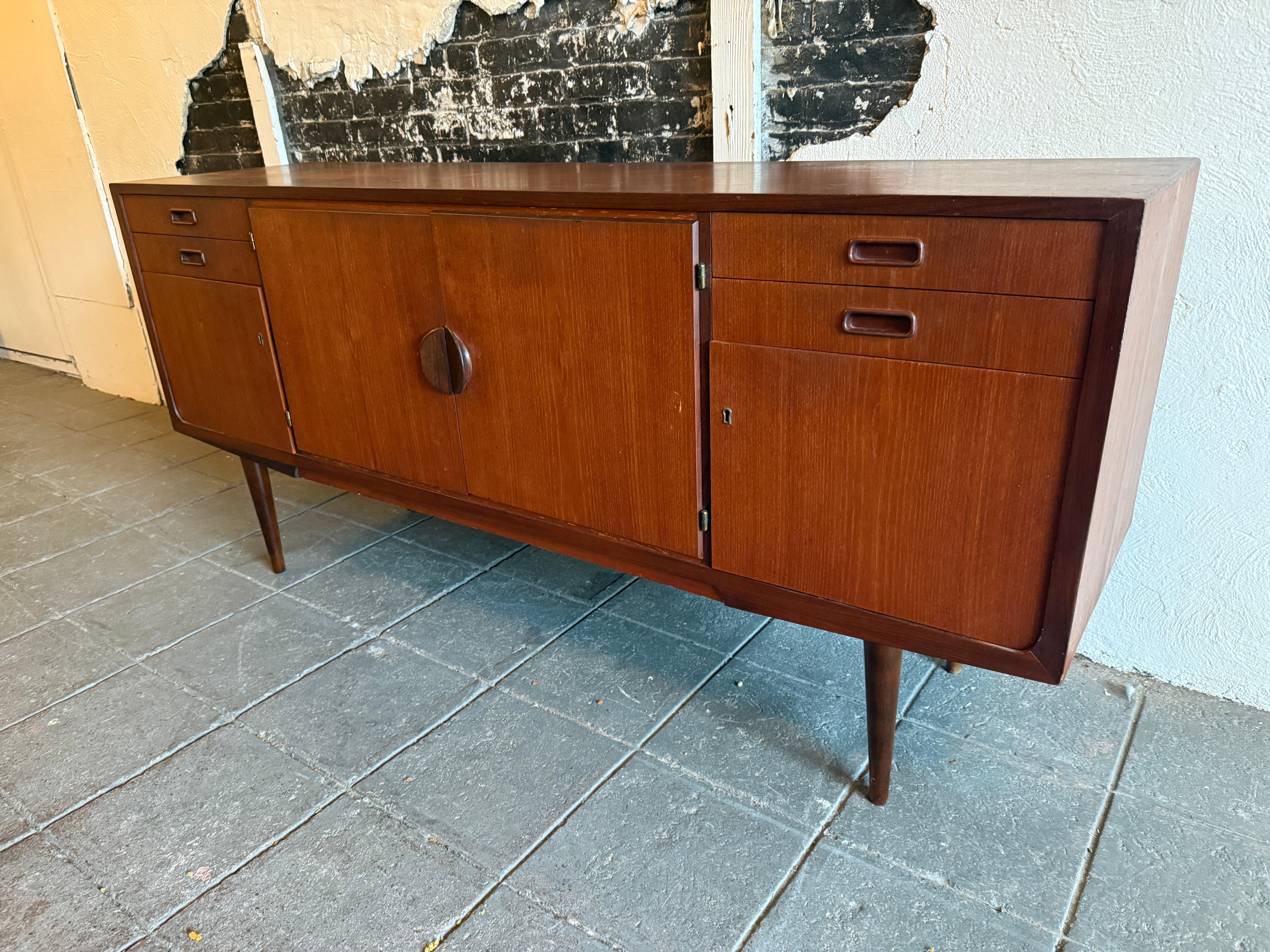 Credenza in teak moderna danese del metà secolo con chiave. Questa credenza in teak presenta (2) ante centrali con maniglie curve e un ripiano centrale dietro di esse. Inoltre ha un design simmetrico con (2) cassetti superiori e (1) cassetto