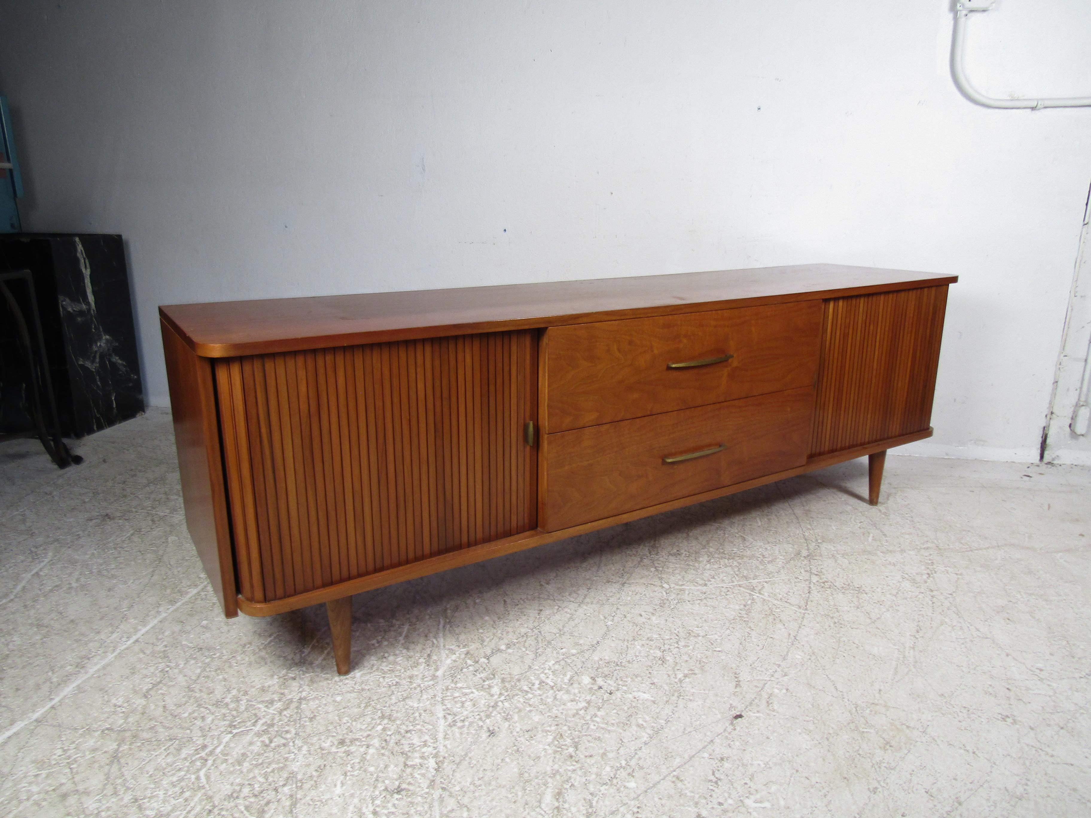 This beautiful vintage modern credenza features tambour doors on each side making this piece stand out from the rest. An elegant walnut finish and four sturdy tapered legs display quality craftsmanship. The large compartments with adjustable shelves