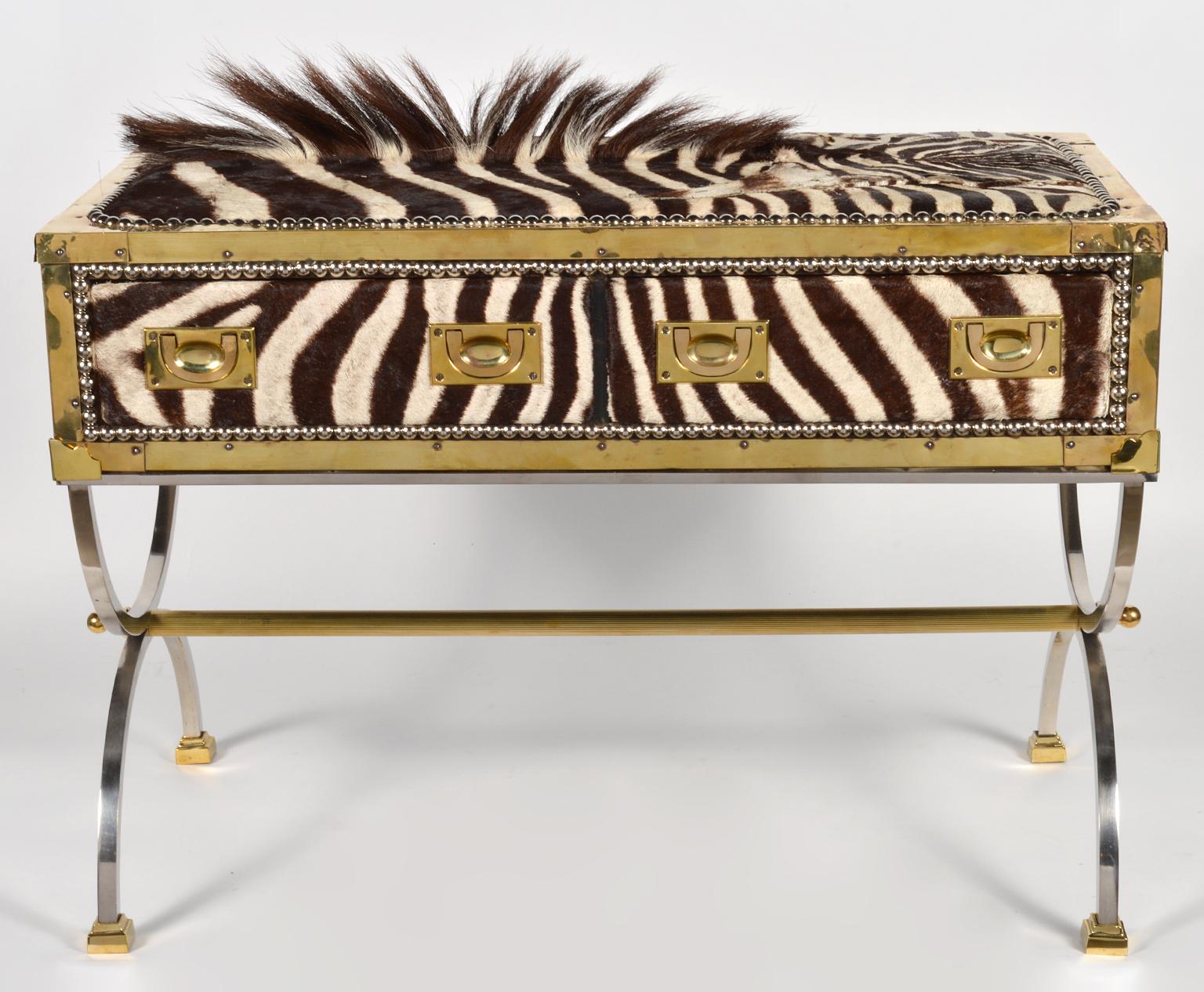 This eye-catching commode features a two drawer brass edged campaign style box or trunk covered with zebra skin on the front and top and with black leather on back and sides accented by steel nail head trim. It rests on a curule style finely