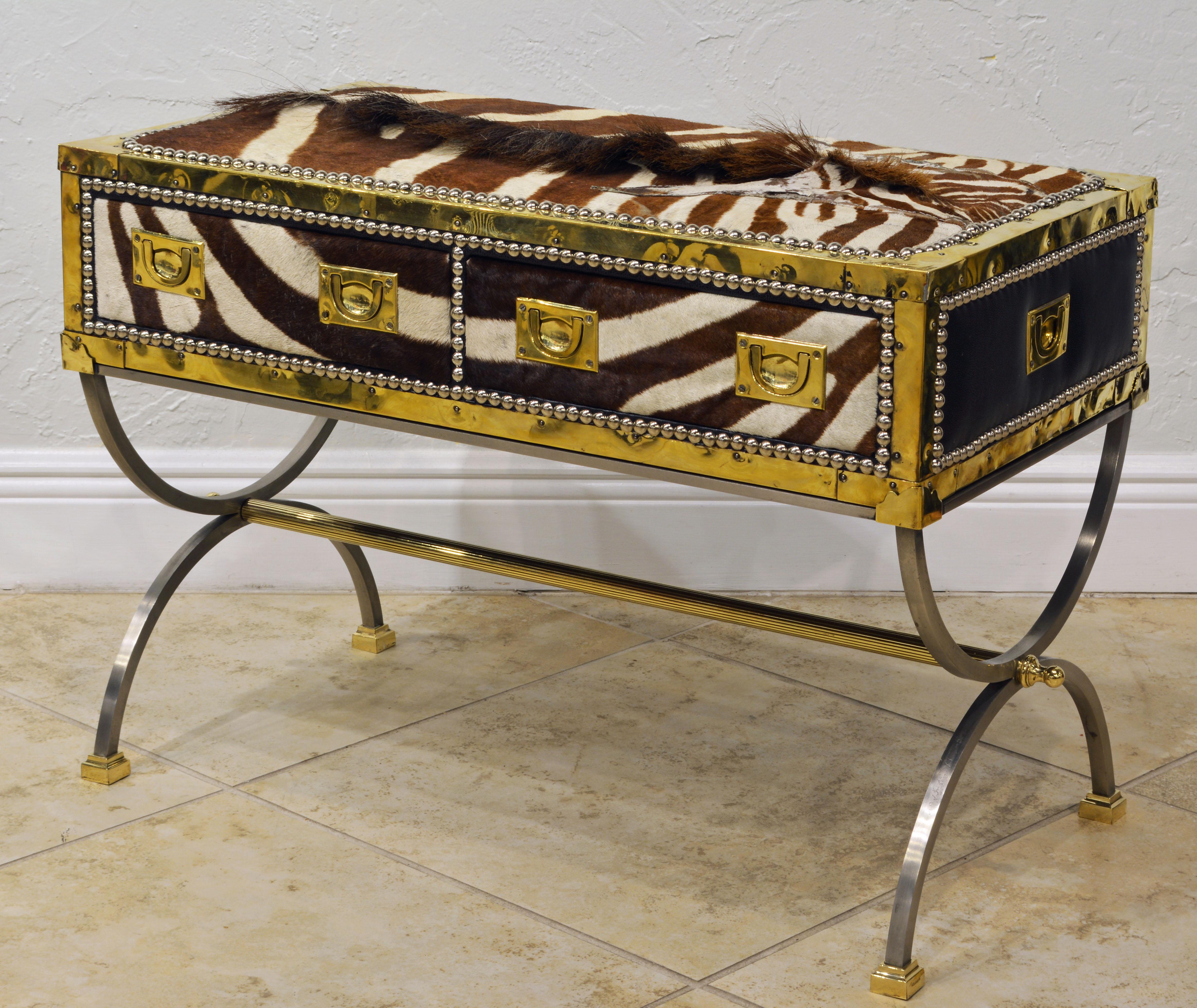 This eye-catching commode features a two-drawer brass edged campaign style box or trunk covered with zebra skin on the front and top and with black leather on back and sides accented by steel nail head trim. It rests on a curule style finely