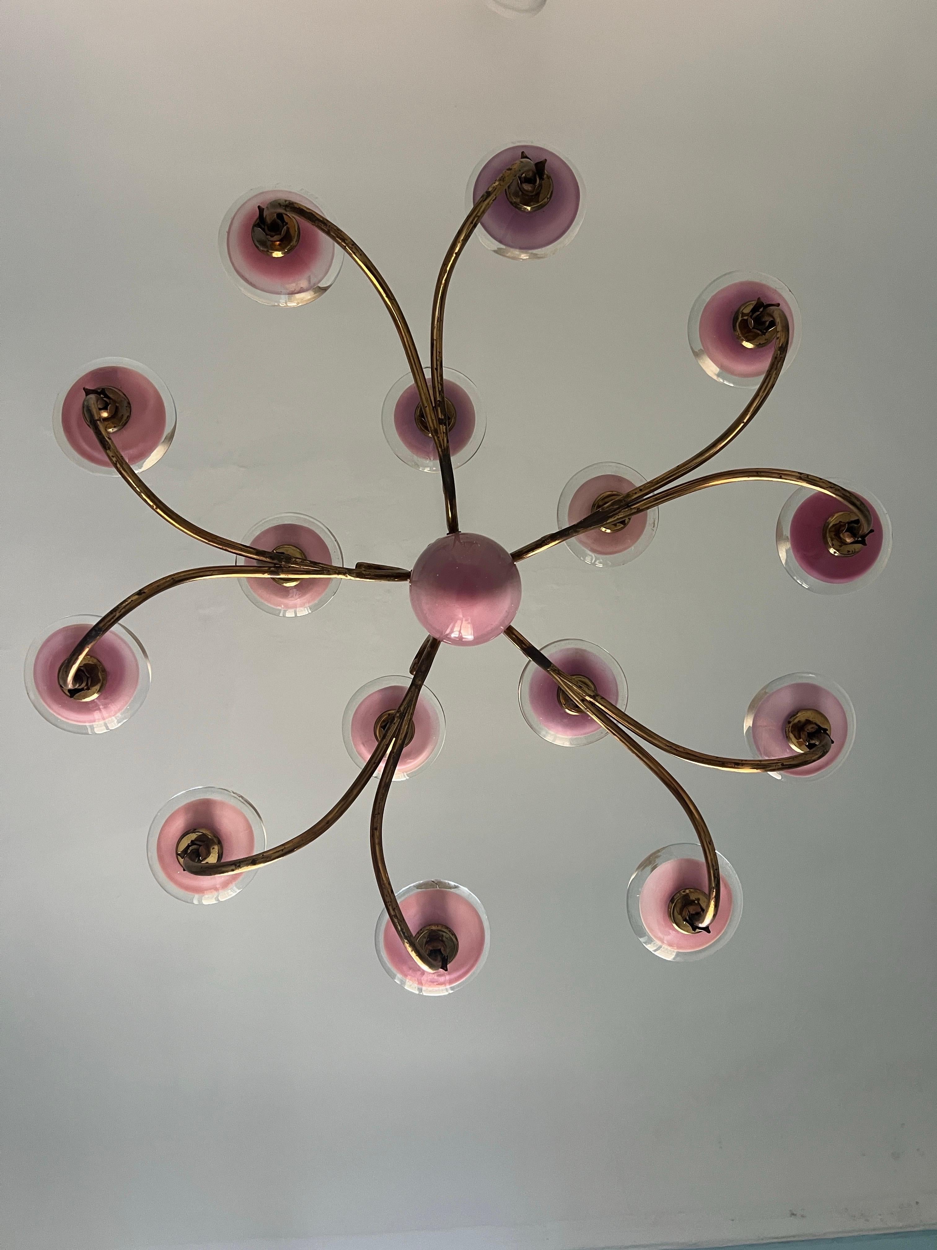 Un lampadario davvero particolare, caratterizzato da eleganti piatti in vetro di Murano rosa incastonati su bracci in ottone che scorrono con grazia. Al centro è appesa una grande goccia di vetro rosa, un punto focale che attira immediatamente lo