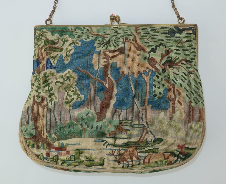 A unique vintage petit point handbag depicting a tranquil forest scene complete with trees, blue skies and deer all in shades of blue, green, brown and gray.  The gold metal frame is detailed with engraving at the top and the kiss lock closure is