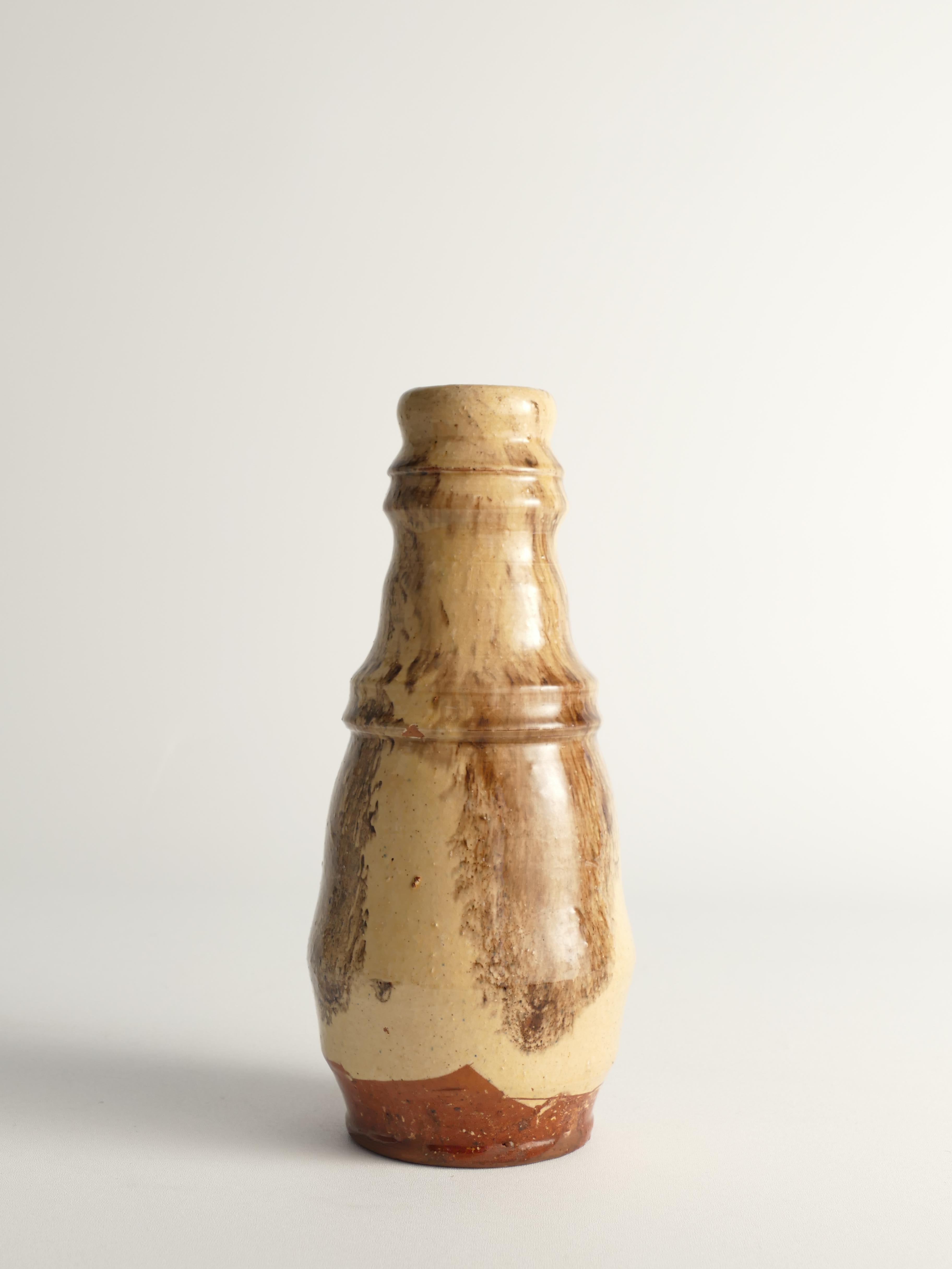 Este singular jarrón moderno escandinavo tiene forma de calabaza, cuerpo bulboso y labio ligeramente acampanado. Está hecho de arcilla rojiza, con la mayor parte del cuerpo cubierto de un vidriado beige cremoso que gotea libremente, acentuado por