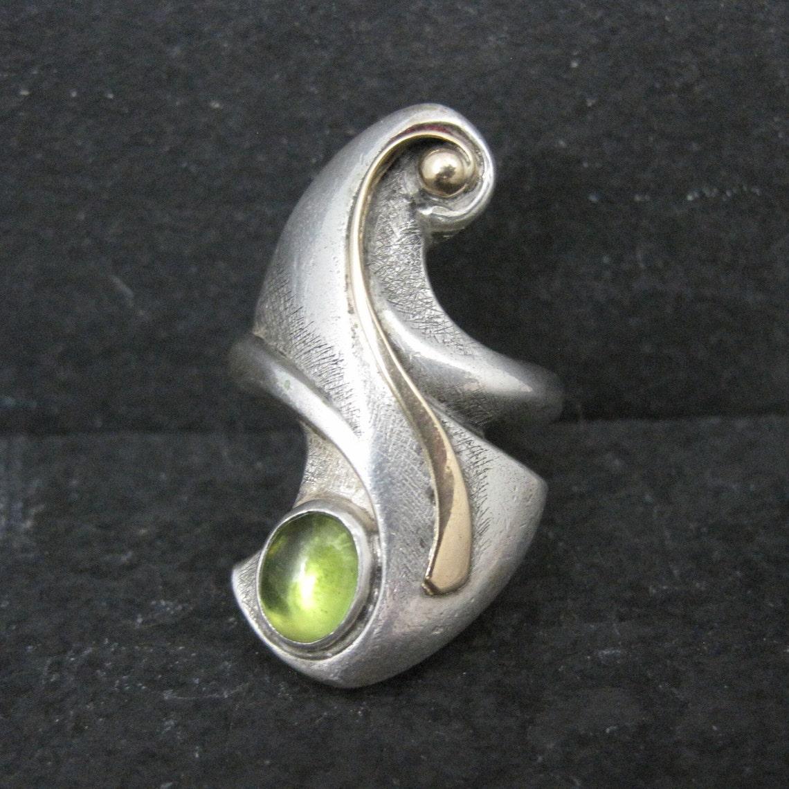 This gorgeous, bohemian style ring is sterling silver and 14k yellow gold with a 6x8mm peridot cabochon gemstone.
It is a product of jewelry artist Patty Graves.

The face of this ring measures 1 3/8 inches north to south.
Weight: 14.8 grams
Size: 8