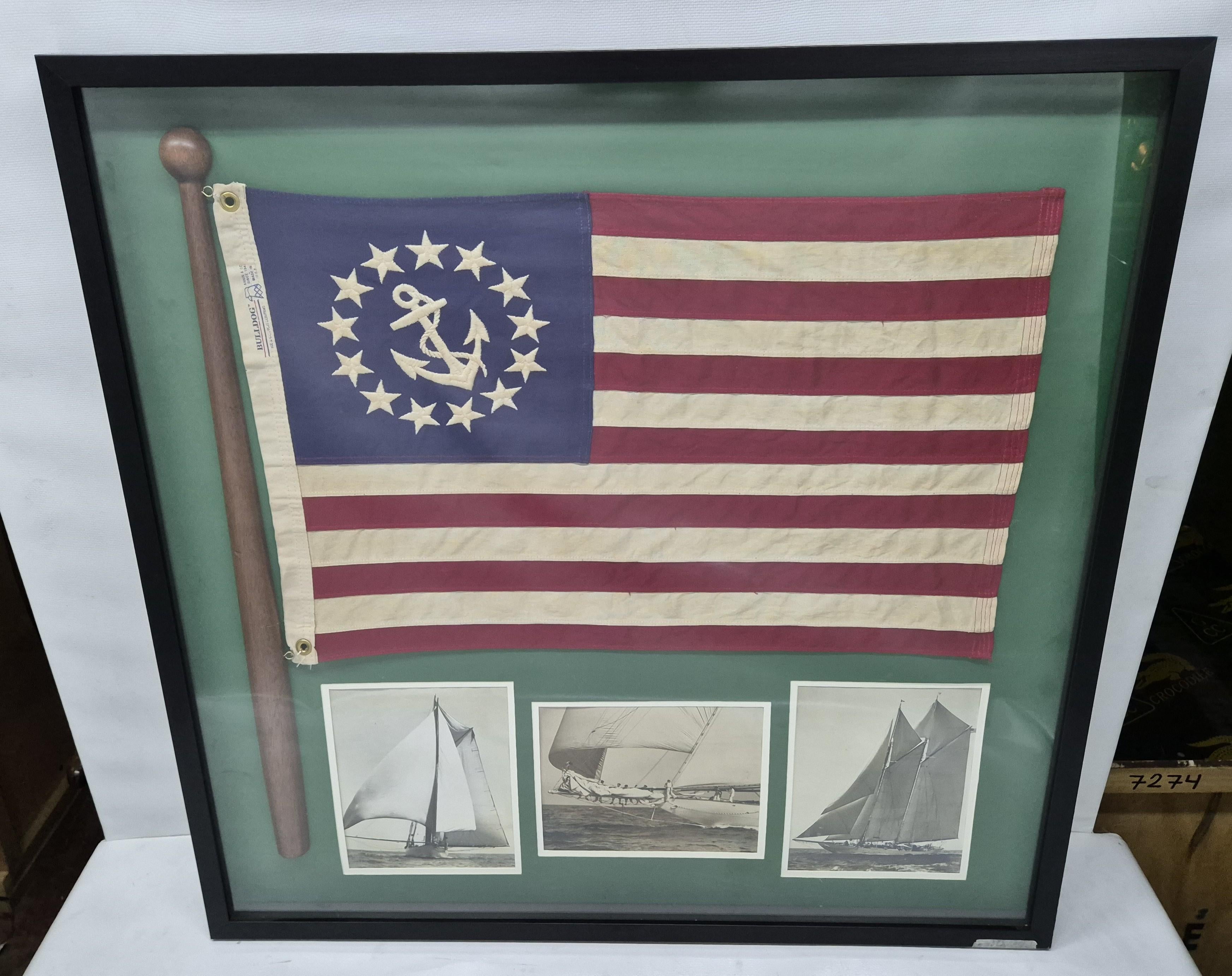 Insignia de yate sobre asta de madera y tres fotografías de yate en blanco y negro montadas en un marco de caja de sombra. La bandera tiene un ancla bordada y trece banderas en el campo azul. La bandera está sujeta a un asta de madera. En el