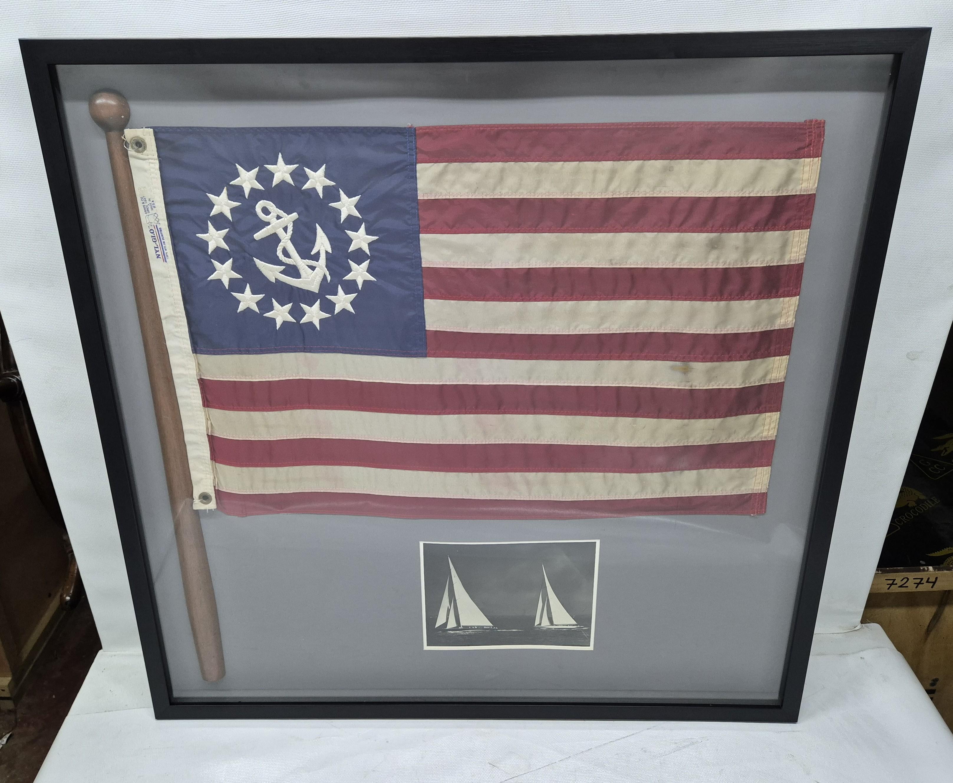 Insignia de yate sobre asta de madera y tres fotografías de yate en blanco y negro montadas en un marco de caja de sombra. La bandera tiene un ancla bordada y trece banderas en el campo azul. La bandera está sujeta a un asta de madera. En el