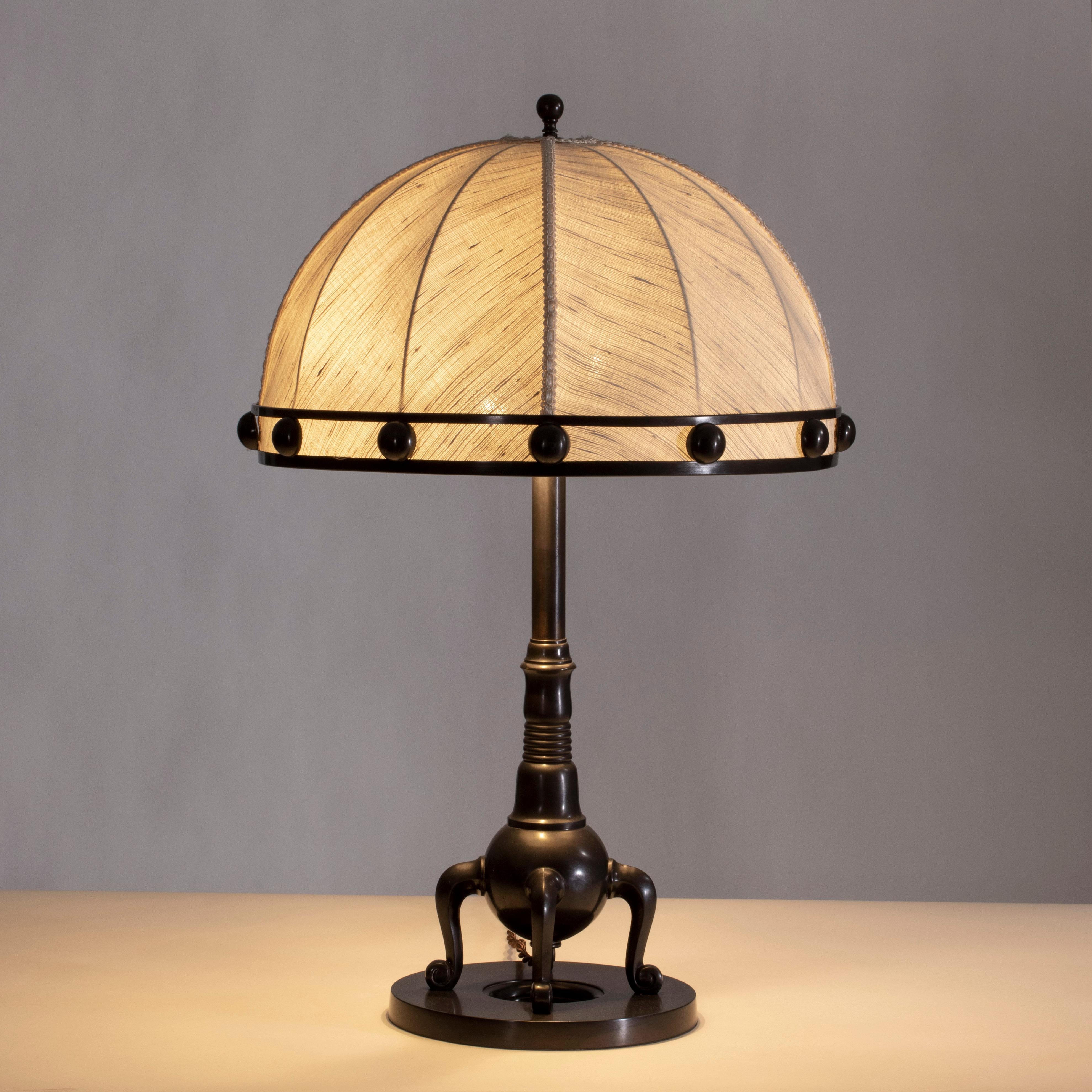 The ball finial, ingeniously doubling as the light switch, above a domed shade mounted with a studded collar in patinated brass, the matching brass standard resting on four s-curved and scrolled legs, on an open circular base. 

Rewired for the