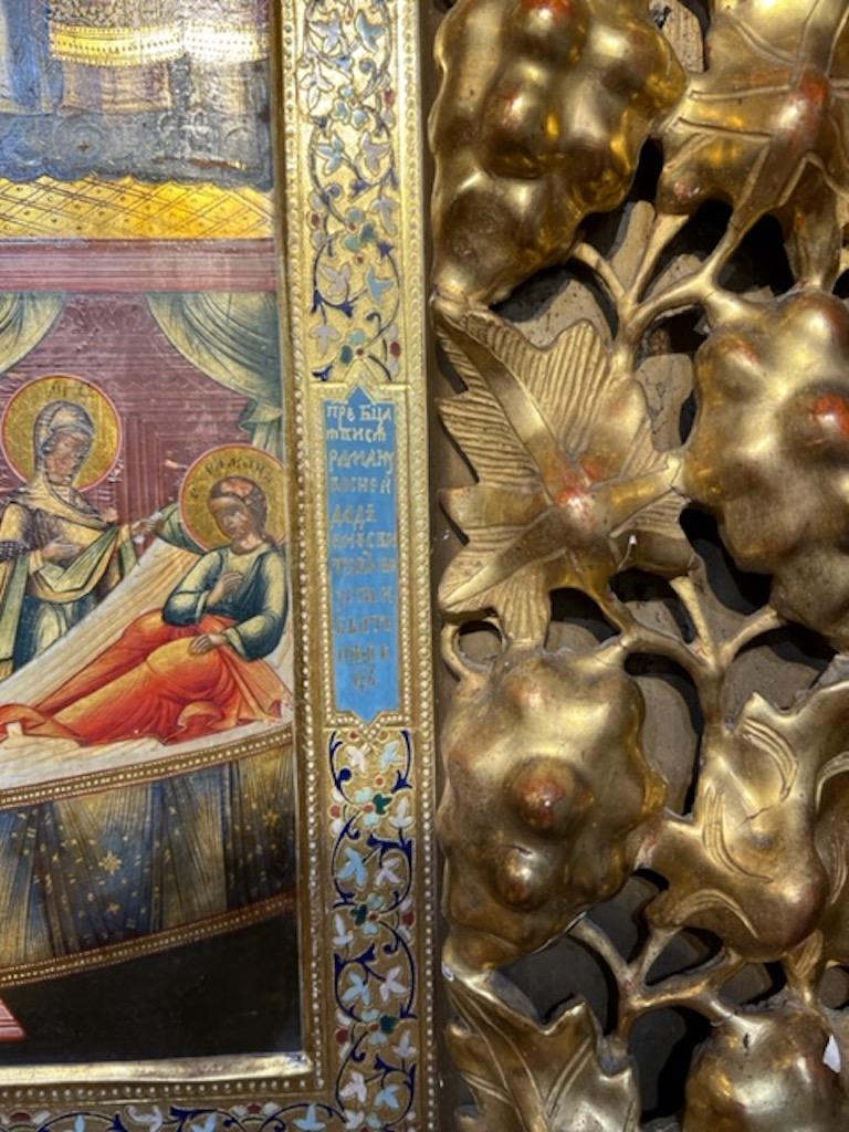 Russische Ikone, Öl auf Holz, mit schönem Cloisonné-Email umrandet. Die Ikone ist von einem dekorativen Einsatz mit Blumenmotiven umgeben, der in den dicken geschnitzten Holzrahmen eingesetzt ist. 
  Die Ikone ist verso mit einer Widmung an die