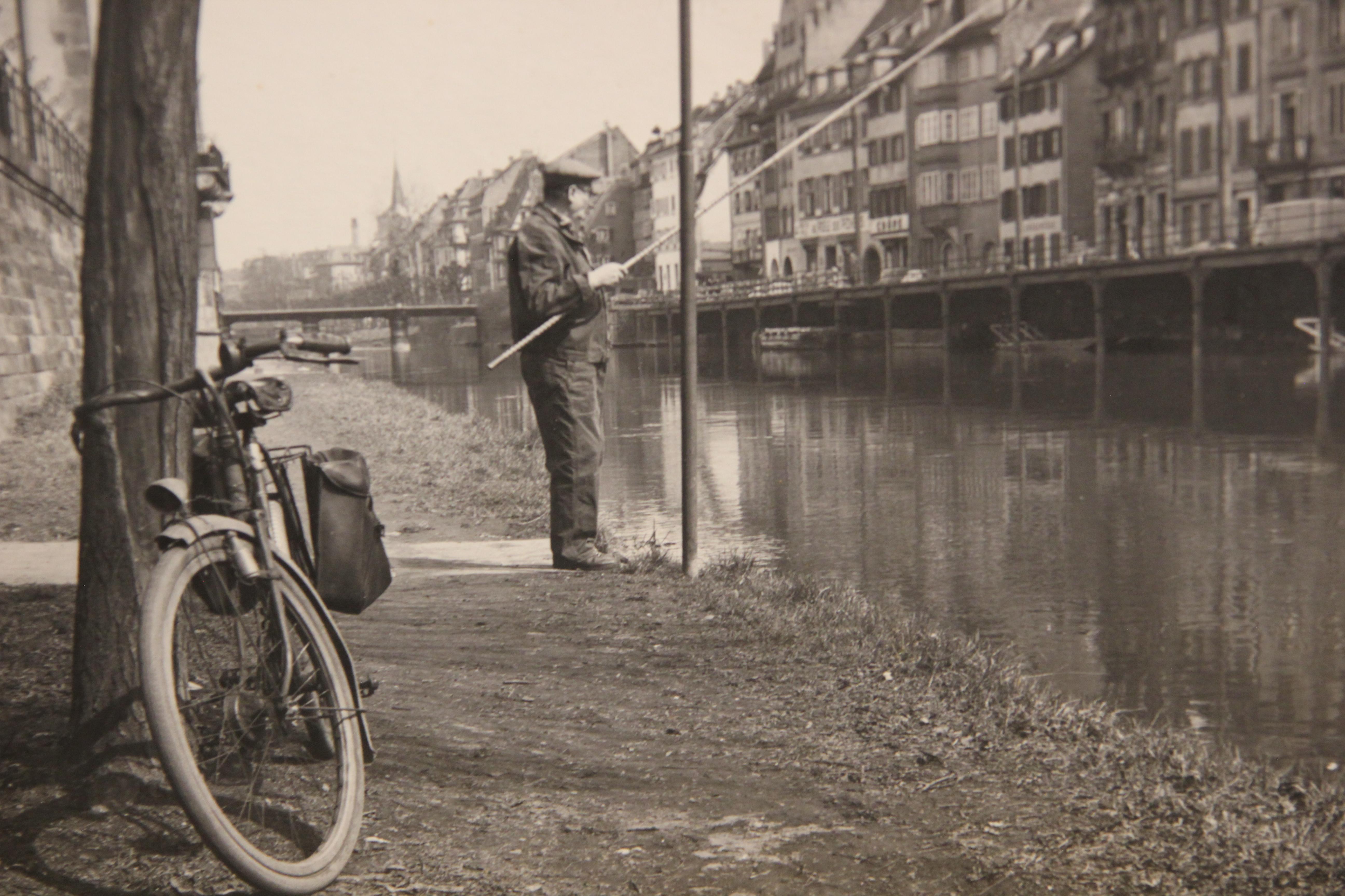 Photographie ancienne en noir et blanc d'un pêcheur qui pêche dans ce qui semble être un décor de ville européenne. La photographie est fixée sur une planche pour éviter toute déformation. La photographie n'est pas encadrée.
 