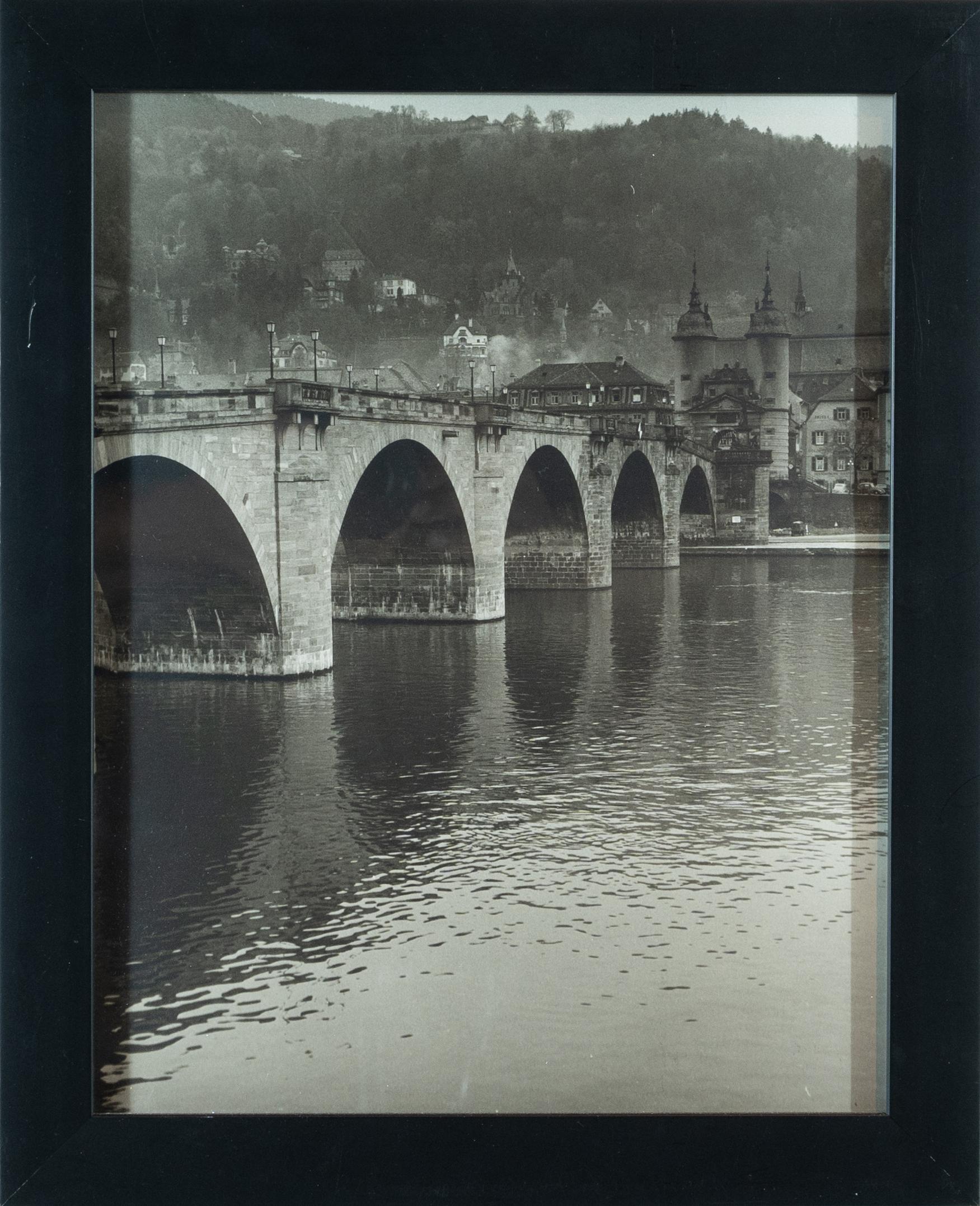 Dieser 20" x 16" große gerahmte Schwarz-Weiß-Fotodruck zeigt eine europäische Brücke, einen Fluss und eine Landschaft. Die Brücke verschwindet auf der linken Seite der Leinwand und führt zu einem scheinbar großen Gebäude auf der rechten Seite des