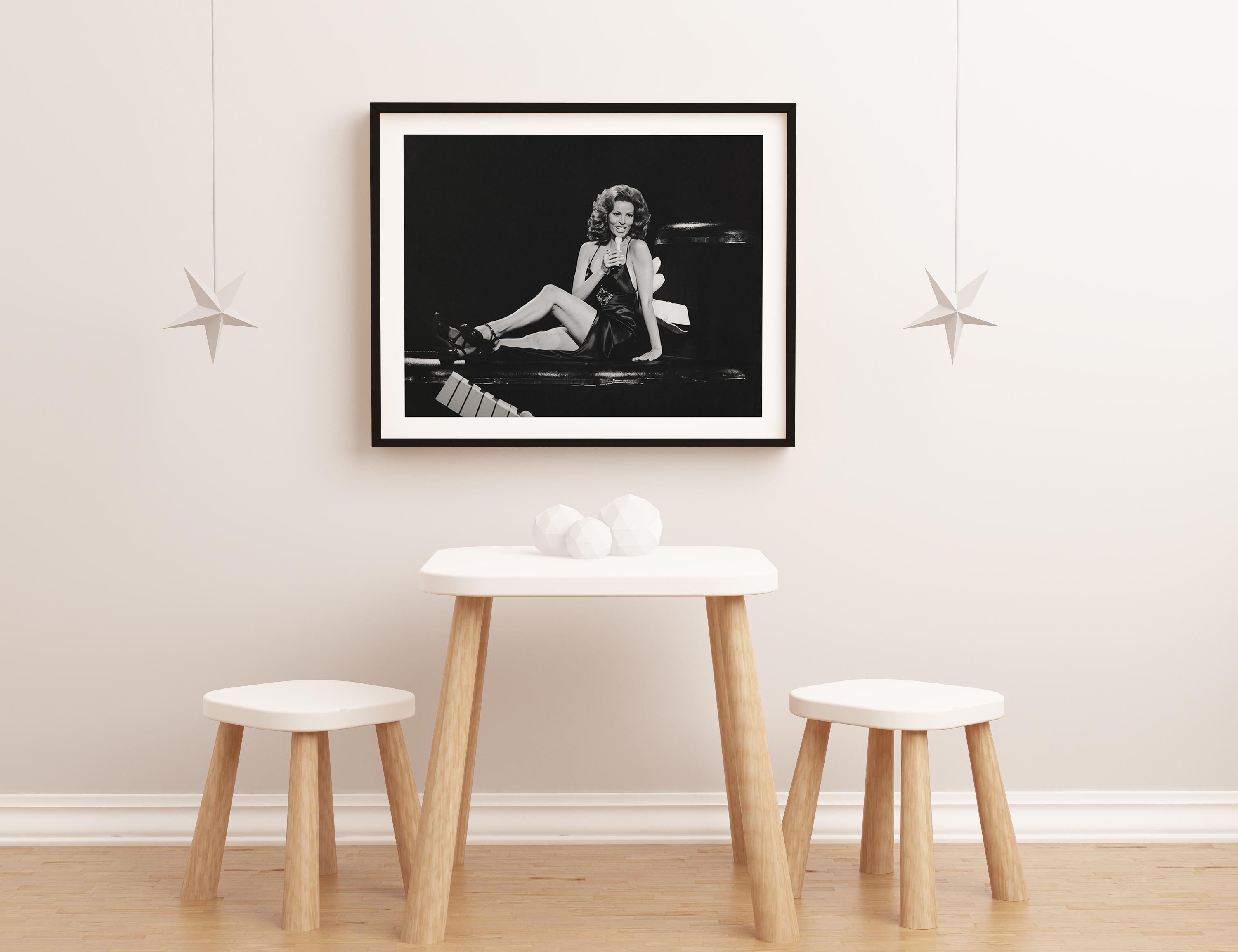 This candid black and white image features Raquel Welch posed seated in a black dress, smiling and singing on stage.

Raquel Welch is an American actress and singer. She first won attention for her role in Fantastic Voyage, after which she won a