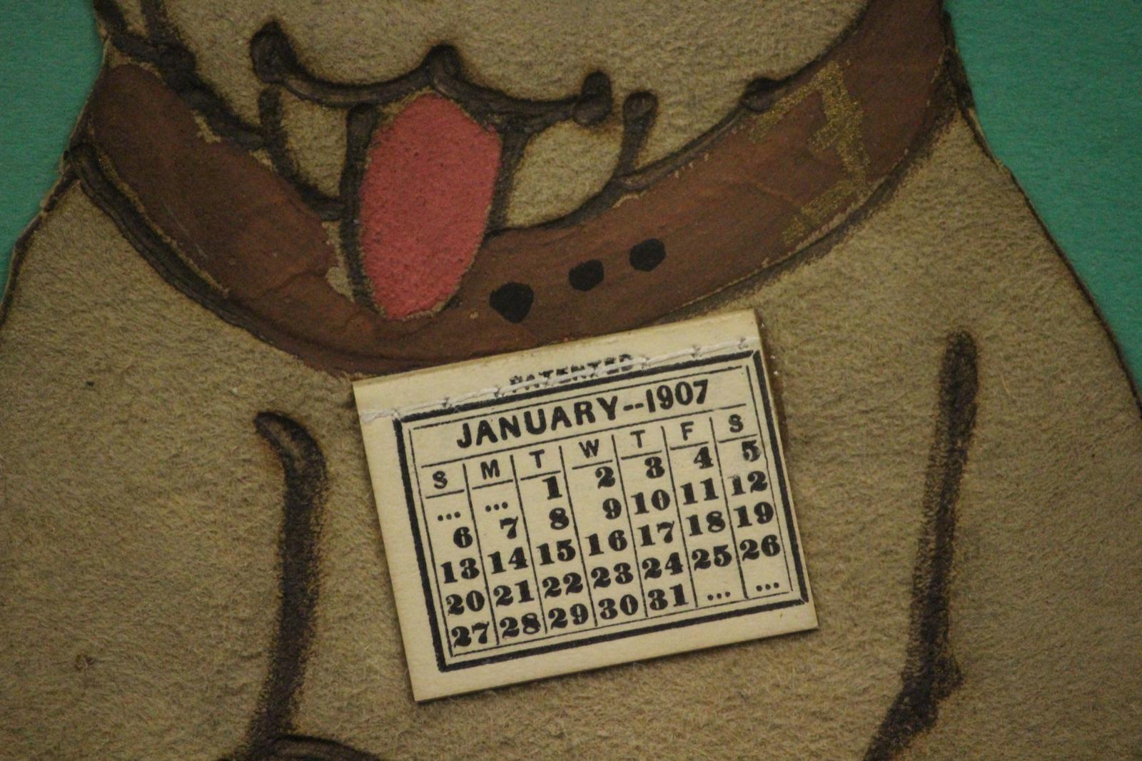 Decoupage-Hund, der eine Geldtasche mit einem am Halsband befestigten Kalender für Januar 1907 umklammert

Bildgröße: 11 5/8 