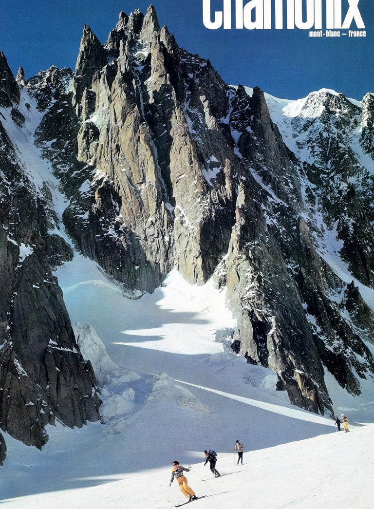 Original vintage ski travel poster for Chamonix Mont Blanc France featuring a great image of skiers skiing in front of a rocky snow covered mountain with the bold white lettering against the blue sky above. The popular resort of Chamonix