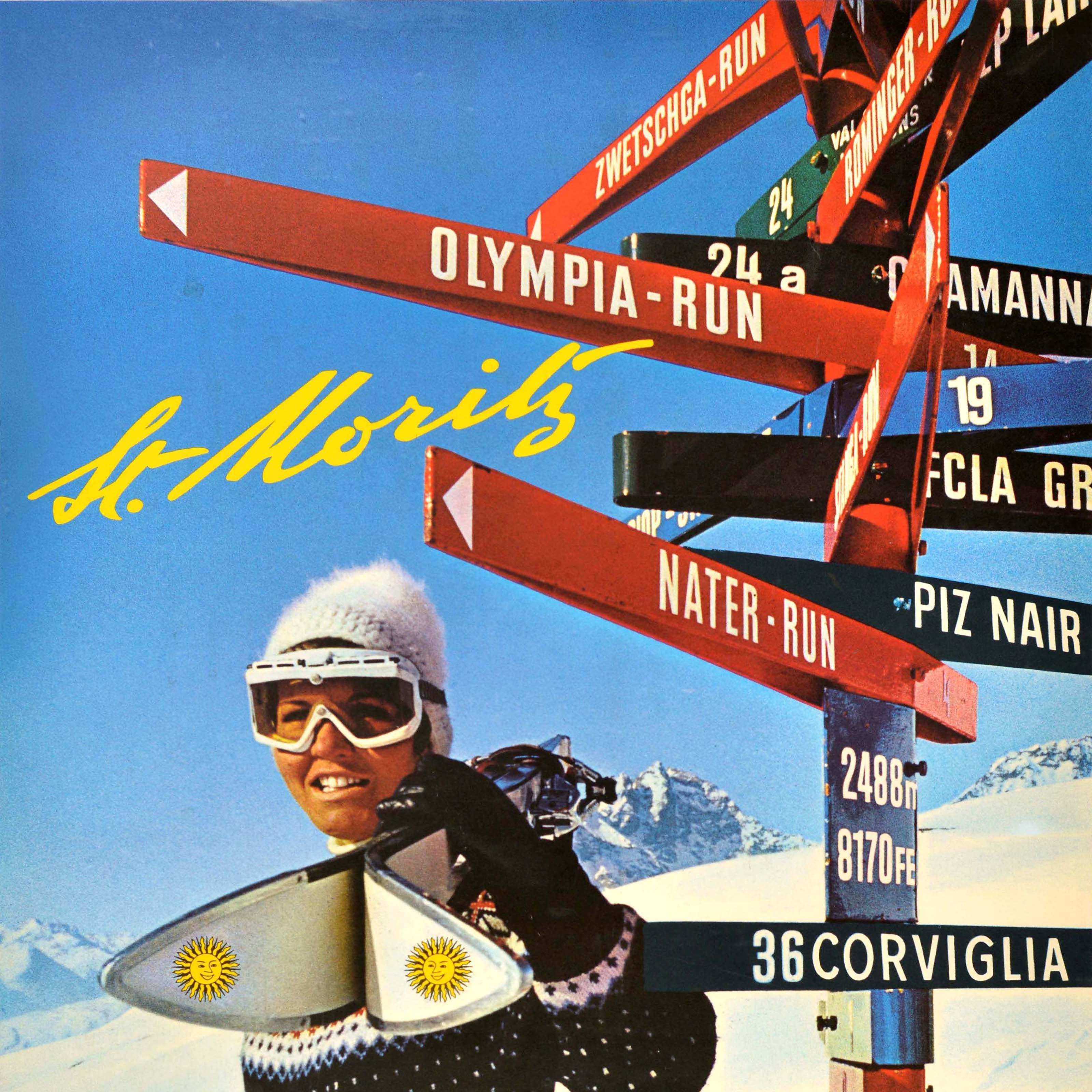 Original vintage skiing poster for the alpine resort of St Moritz in Switzerland featuring a skier wearing ski goggles, a bobble hat and knitted jumper, and holding poles and a pair of skis with the St Moritz sun logo on them, standing in front of a