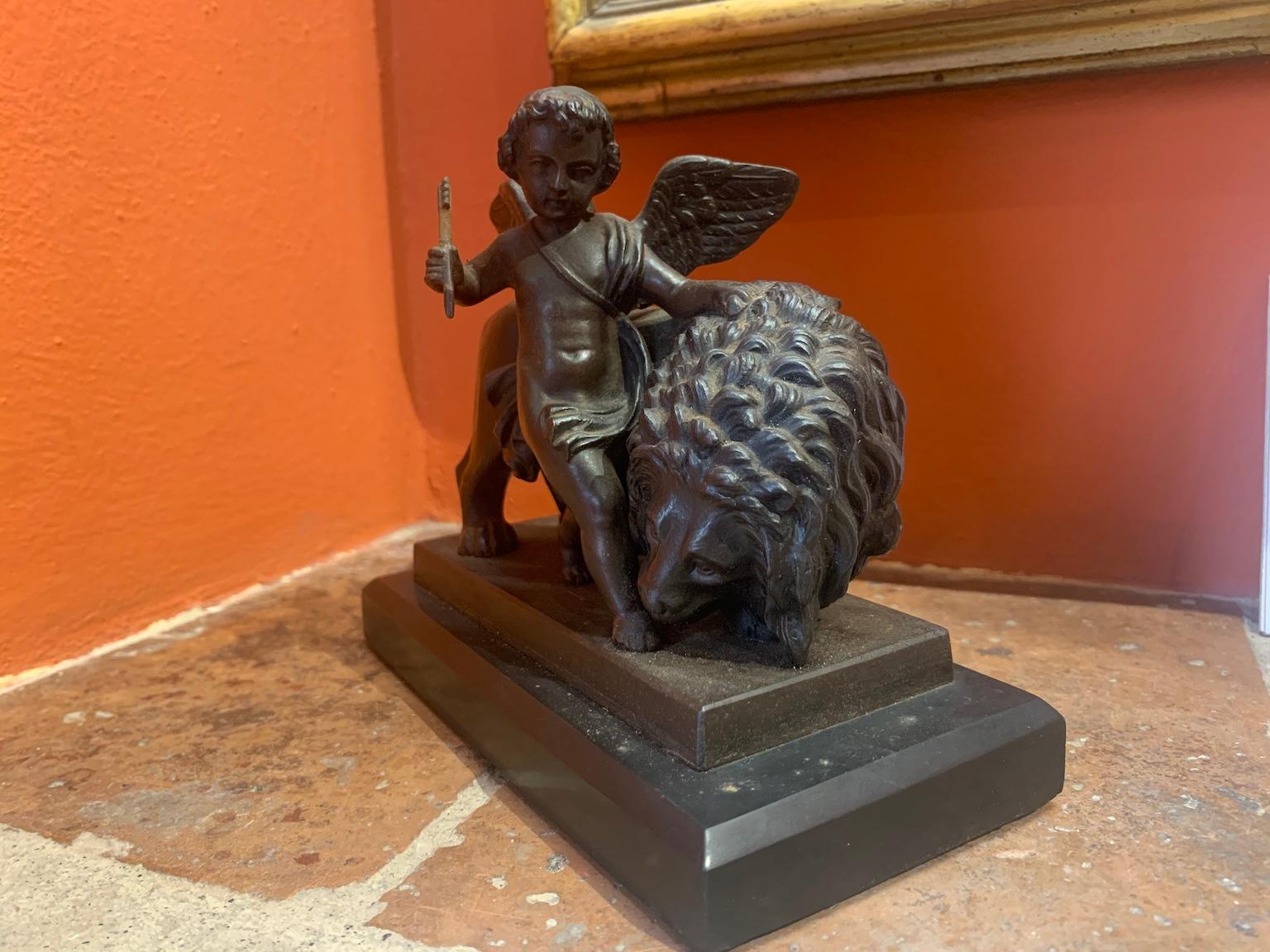 Marble-based bronze statuette depicting Cupid, holding an arrow in his hand, walking caressing a lion's mane, lowered in the act of affectionately licking his little foot. The subject is an allegory of love conquering all, dominating and defeating