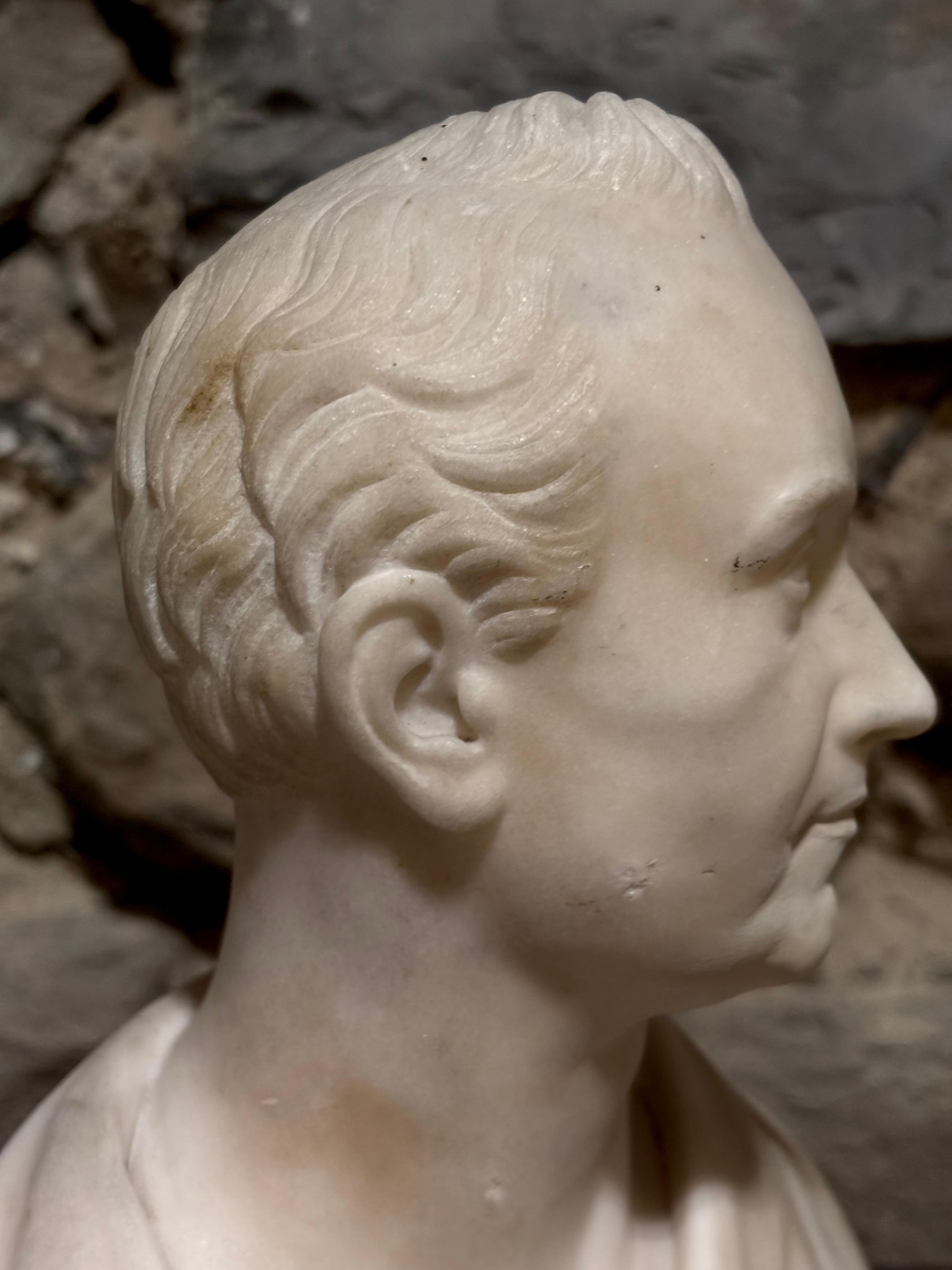 Busto neoclassico in marmo raffigurante un dignitario romano in  erma. Epoca Impero, primo quarto del XIX secolo.   
Si tratta di un piccolo busto da scrivania raffigurante un giovane dignitario con indosso una tunica.  Questi piccoli busti erano