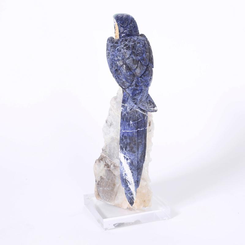 Pappagallo brasiliano scolpito in pietre semipreziose appollaiato su un geode di quarzo e presentato su una base di lucite.