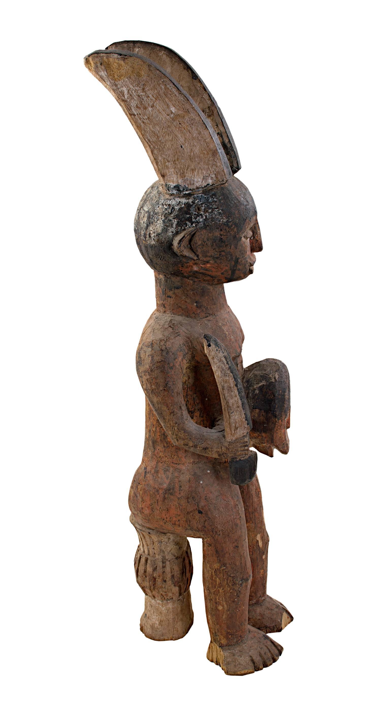 Cette sculpture fétiche Ekamka a été sculptée dans du bois par un artiste nigérian inconnu. Elle représente un personnage tenant un masque. Le personnage a deux cornes symboliques qui dépassent de sa tête et il est assis sur un mince