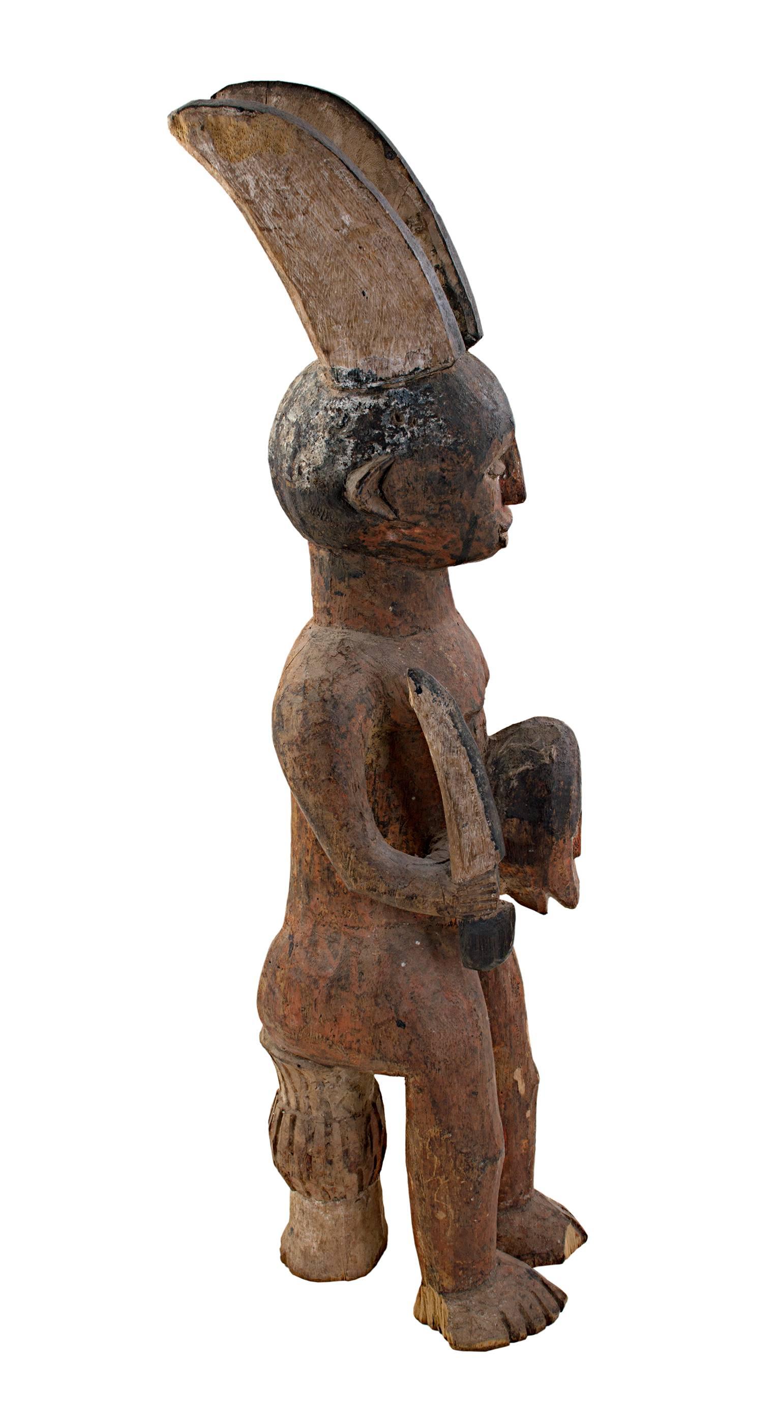 Esta escultura Ekamka Fetiche fue tallada en madera por un artista nigeriano desconocido. Representa una figura que sostiene una máscara. La figura tiene dos cuernos simbólicos que sobresalen de su cabeza y está sentada en un delgado