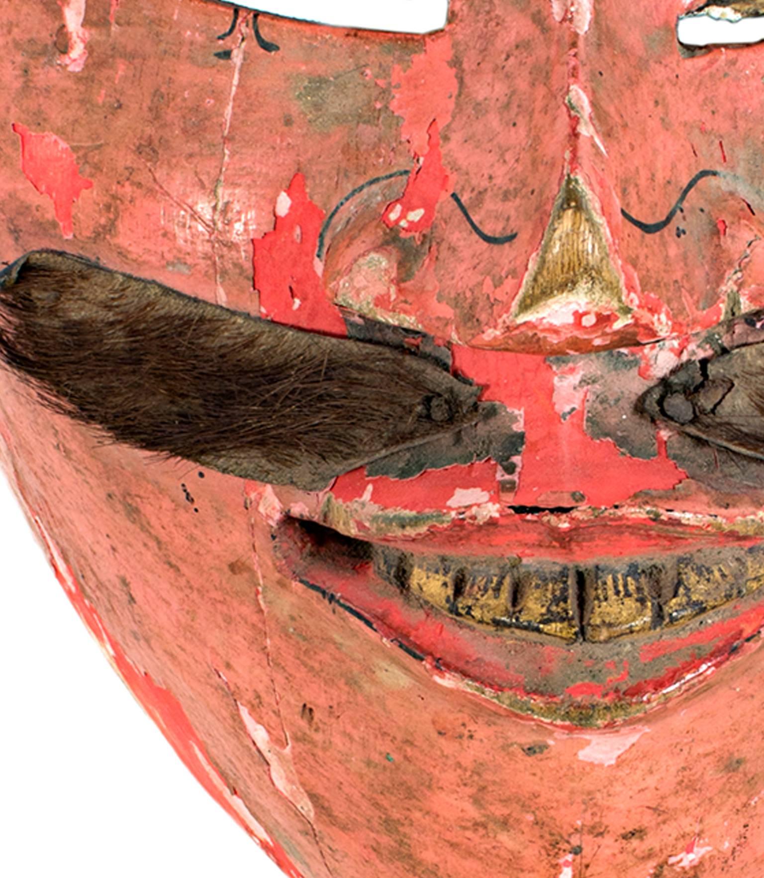 This mask, created by an unknown Indonesian artist, features a bright salmon-colored face with heavily slanted eyes. It also has a mustache made of animal fur. It is approximately 7