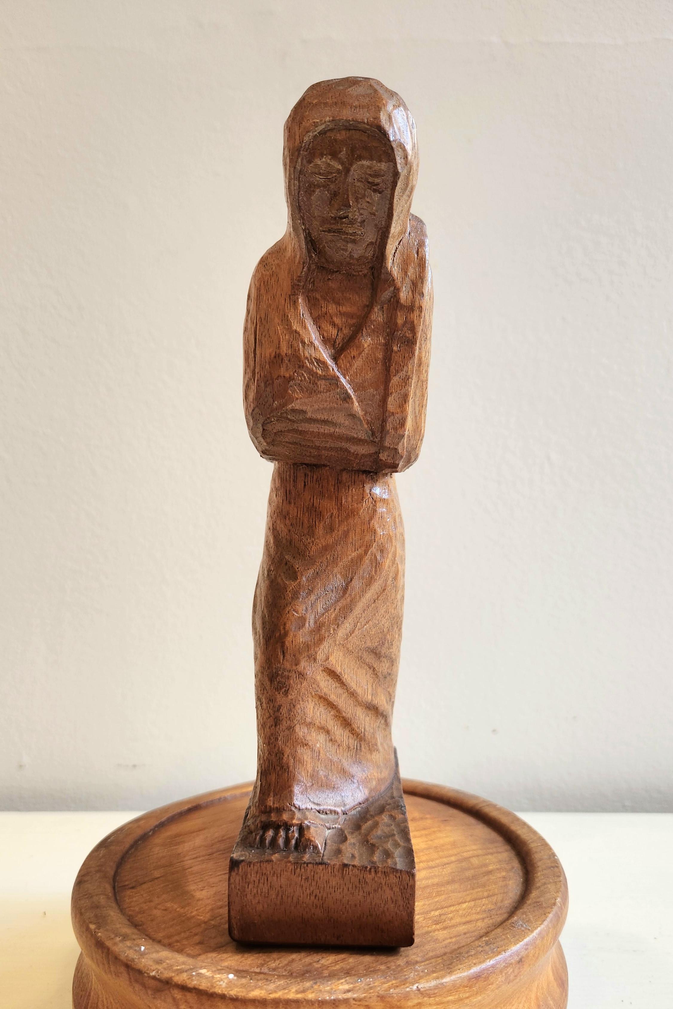 This beautiful wooden figurine depicts a woman in a long robe with a hood  covering her head and carrying a sack over her shoulder. The woman in the sculpture could be viewed as a working woman given the intensity of her face. Most likely, however,