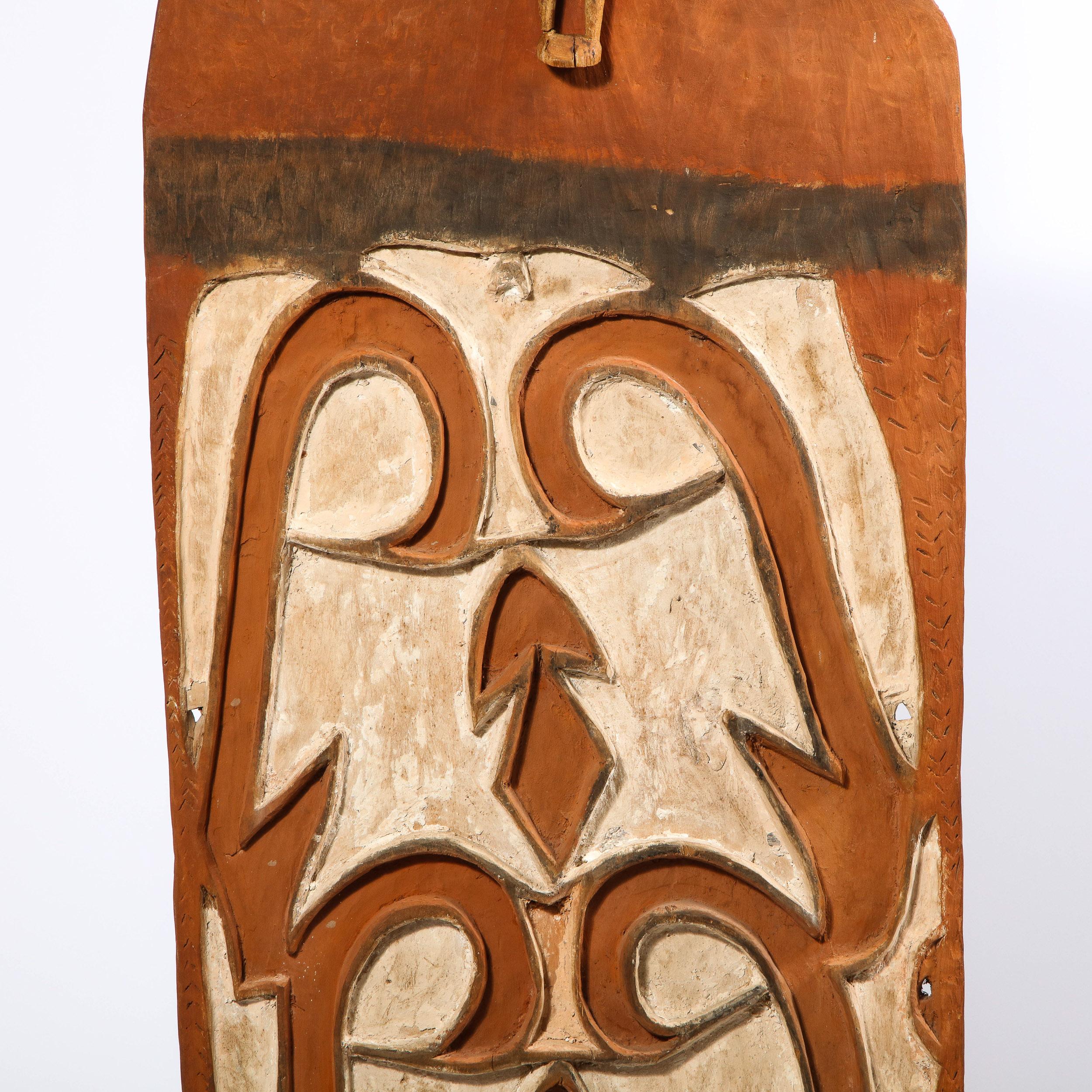 Dieses markante Schild wurde um 1960 in Papua-Neuguinea hergestellt. Die rechteckige Form aus unbehandeltem, nubukfarbenem Hartholz ist mit weißen, geschwungenen und geometrischen Mustern beschriftet. Eine kleine geschnitzte Holzfigur ziert den