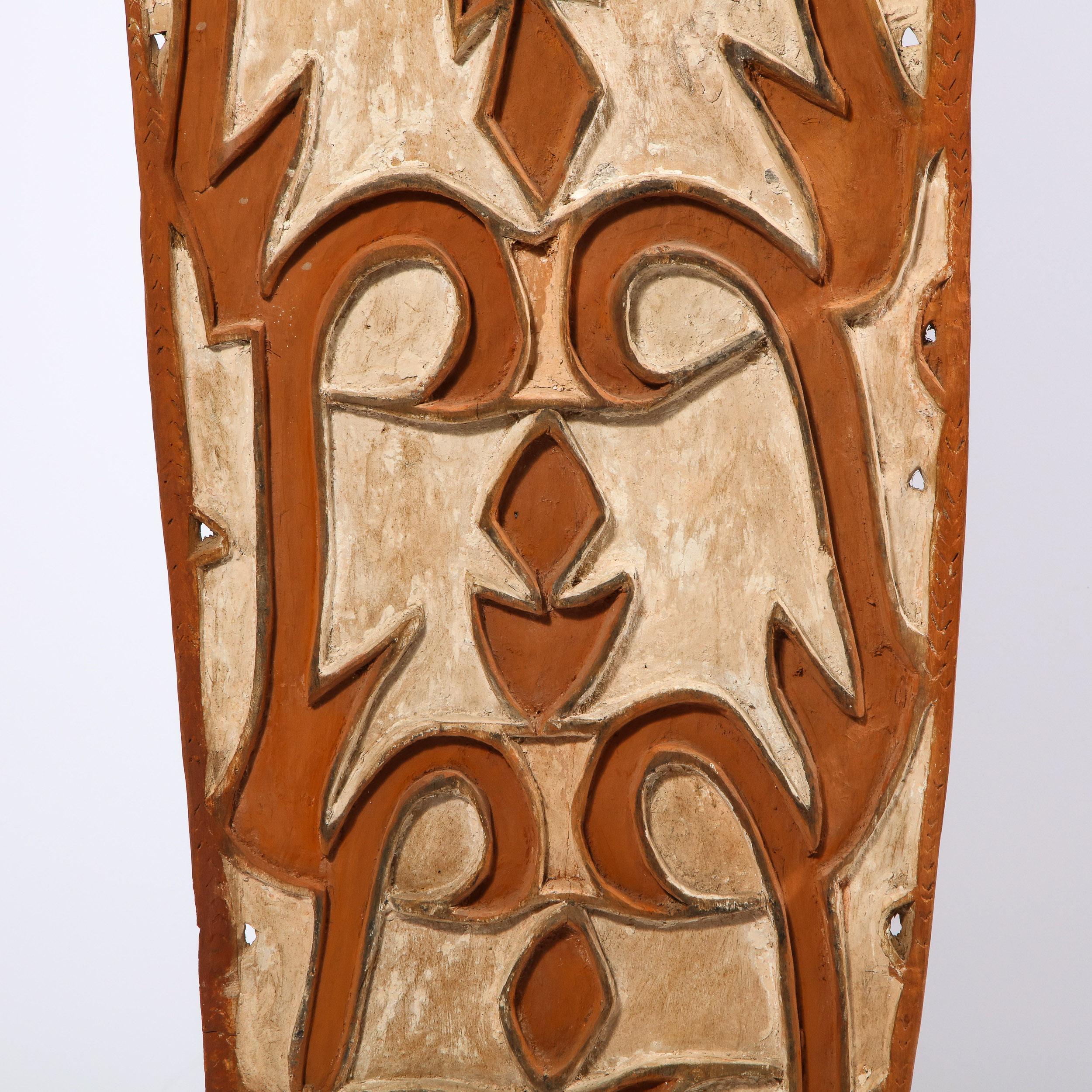 This striking shield was realized in Papua New Guinea, circa 1960. The rectangular form in unfinished nubuck hued hardwood has been inscribed with white curvilinear and geometric patterns. A small carved wood figurine adorns the top of the shield.